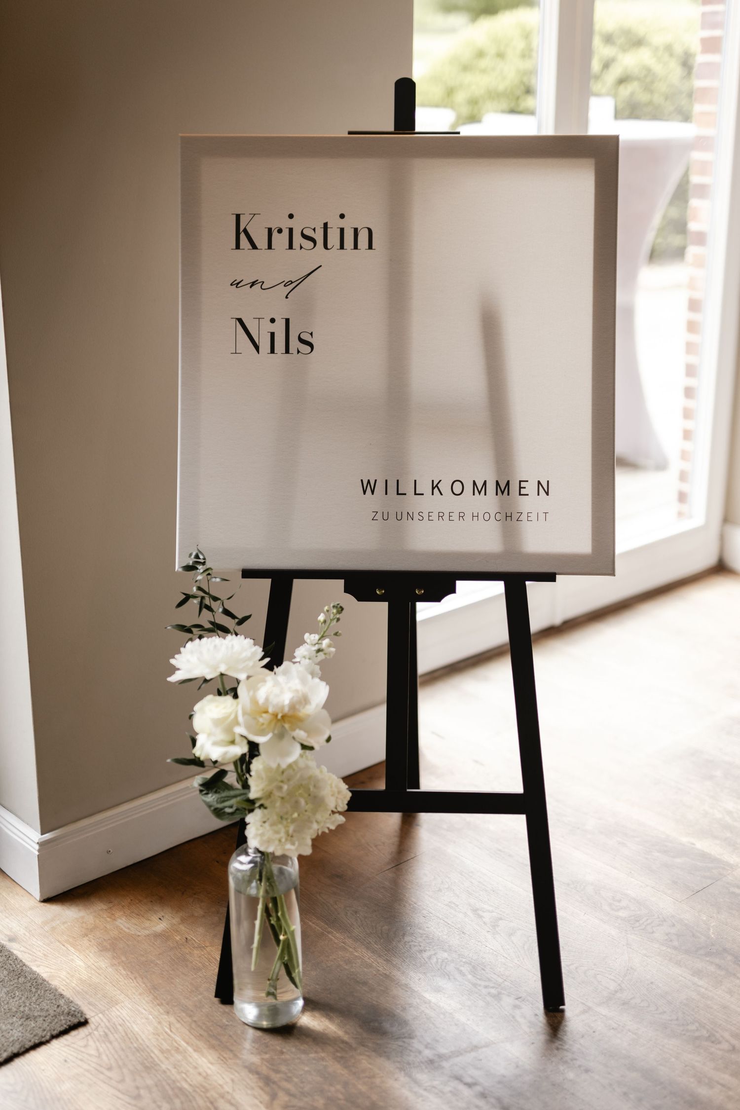 Willkommensschild für Kristin und Nils auf einer Staffelei neben einer Vase mit weißen Pfingstrosen und grünem Blattwerk.  Scheune St. Georg