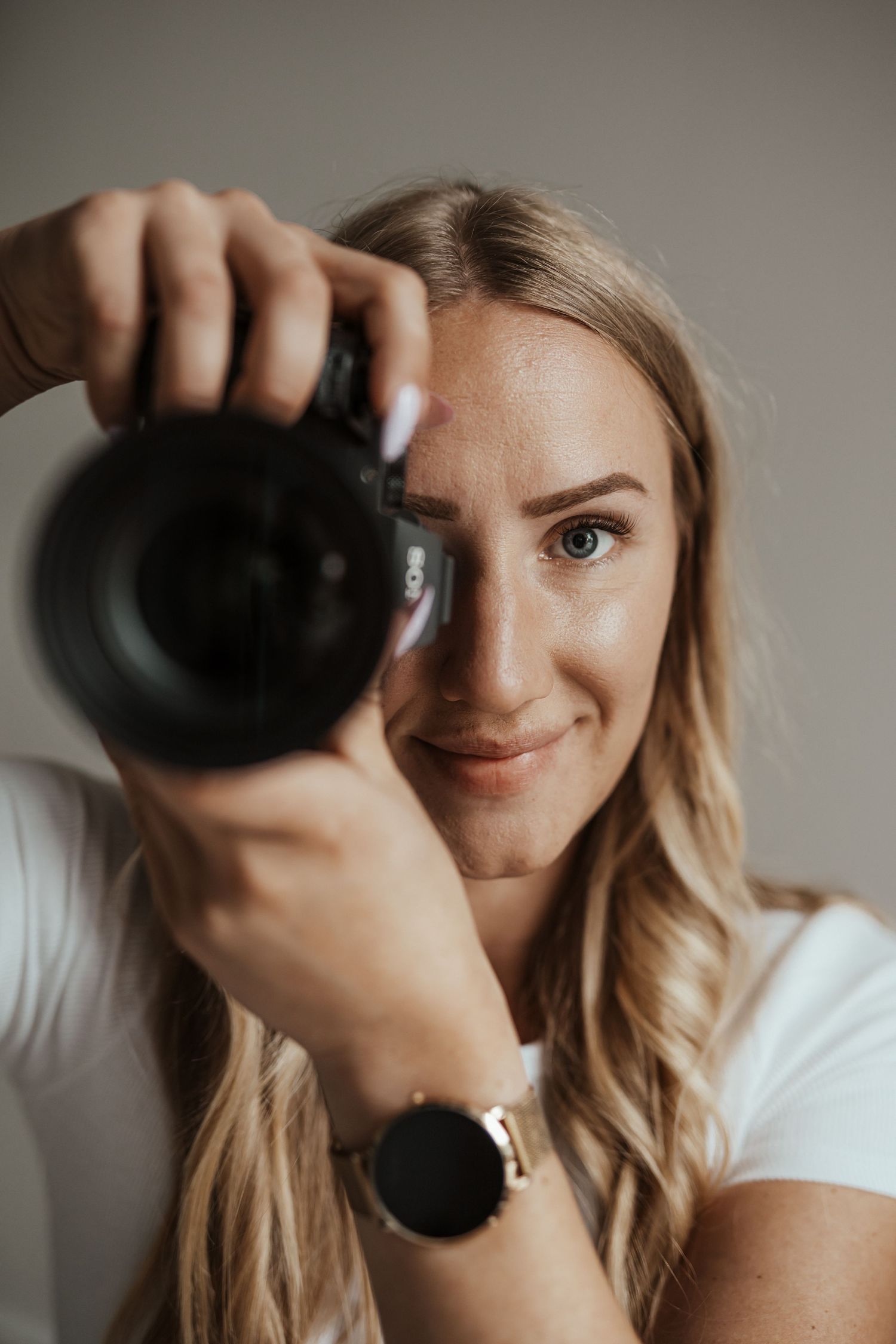 Fotografin mit welligem blondem Haar hält ihre Kamera vor das Gesicht, sodass ein Auge sichtbar bleibt.  Sarah Appel Fotografie