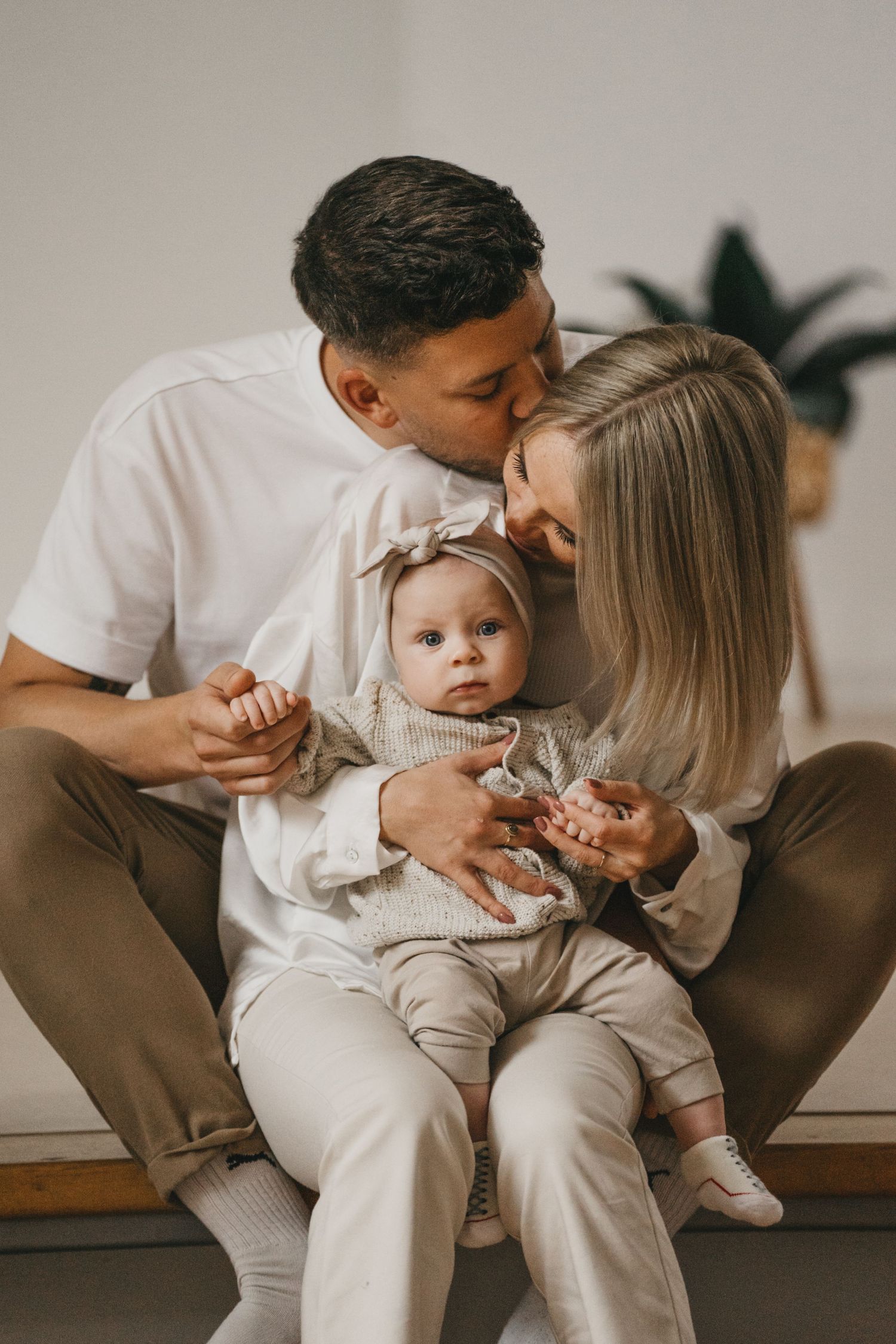Parents sitting together kissing and holding their baby girl in a headband in a bright, cozy family portrait.