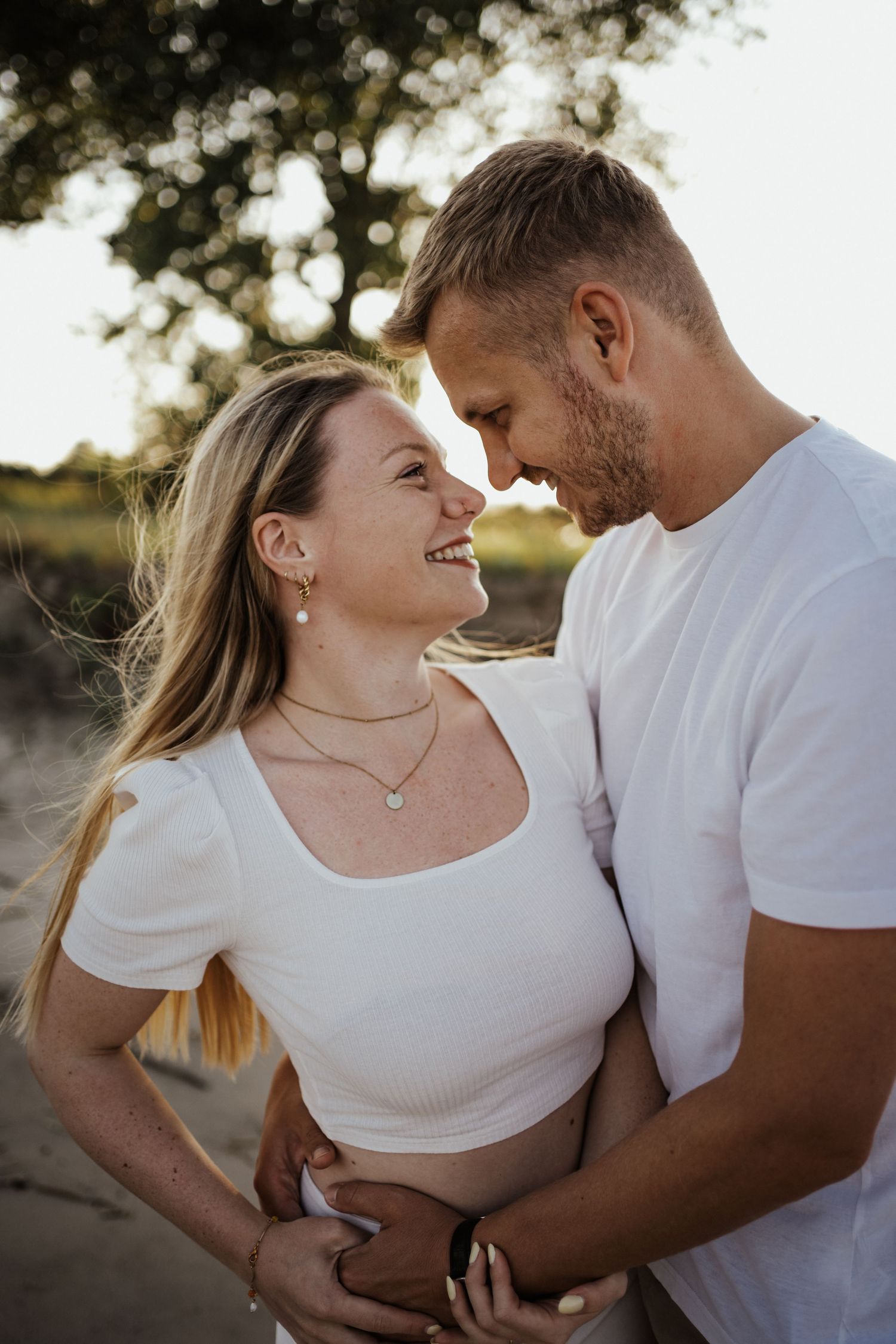 A smiling couple shares an affectionate moment during an outdoor engagement photoshoot in soft golden sunlight.