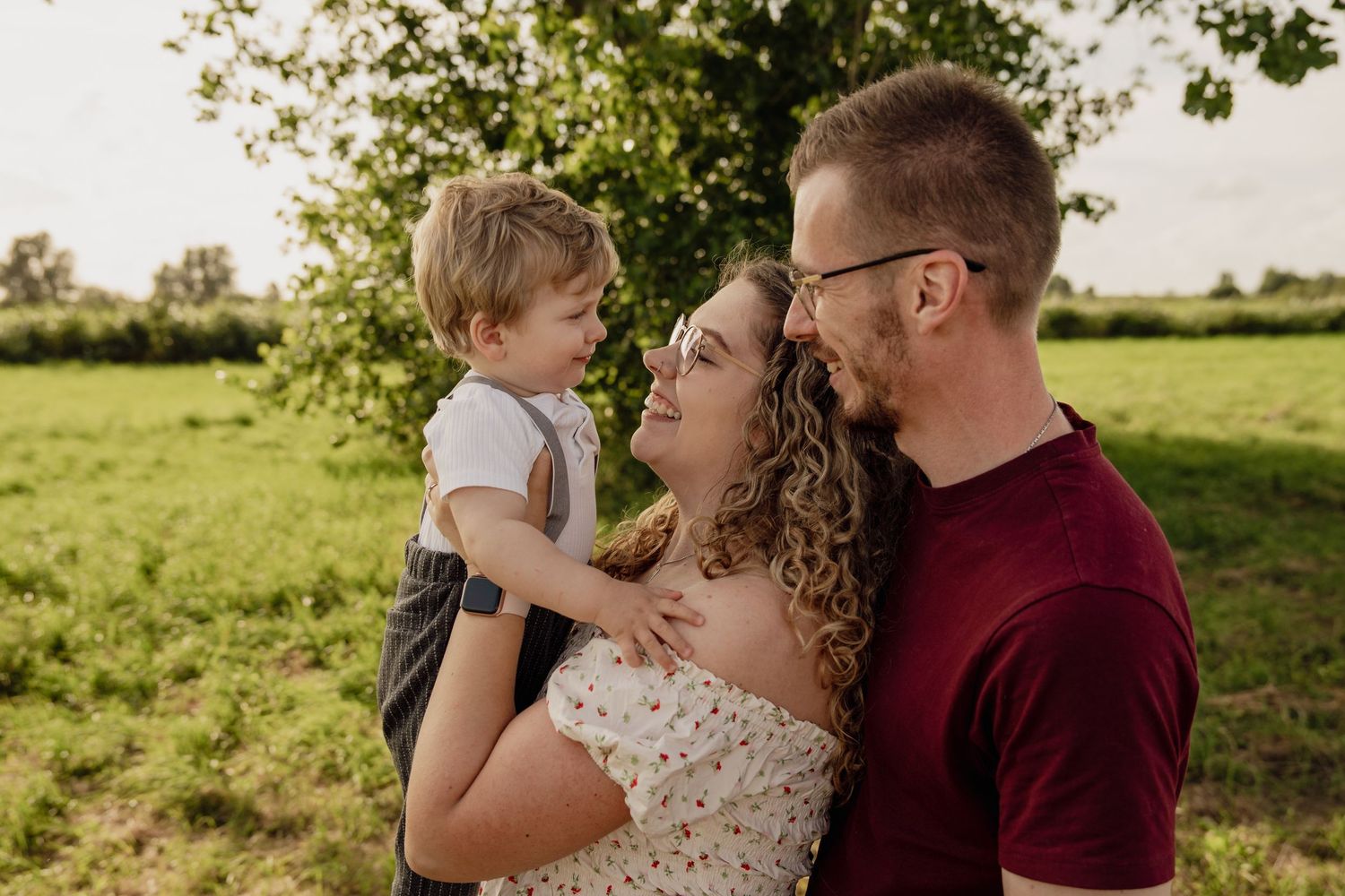 Lächelnde Eltern halten ihren kleinen Sohn gemeinsam zwischen sich auf einer grünen Wiese beim Familienfotoshooting.