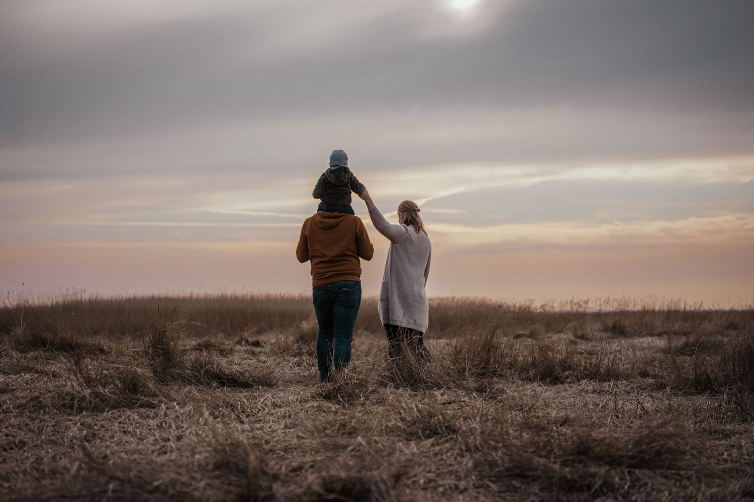 Family silhouette walking through a grassy field at sunset, with a child riding on a parent's shoulders under a soft sky.