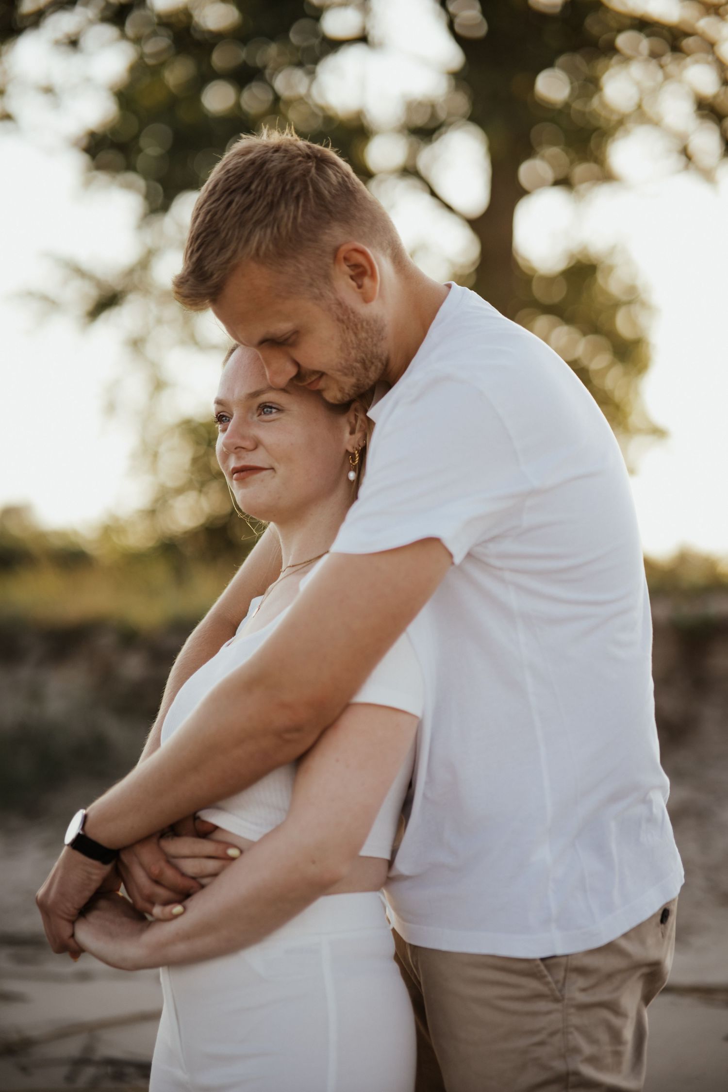 A man tenderly embraces a woman from behind and rests his face against her head in warm evening sunlight.