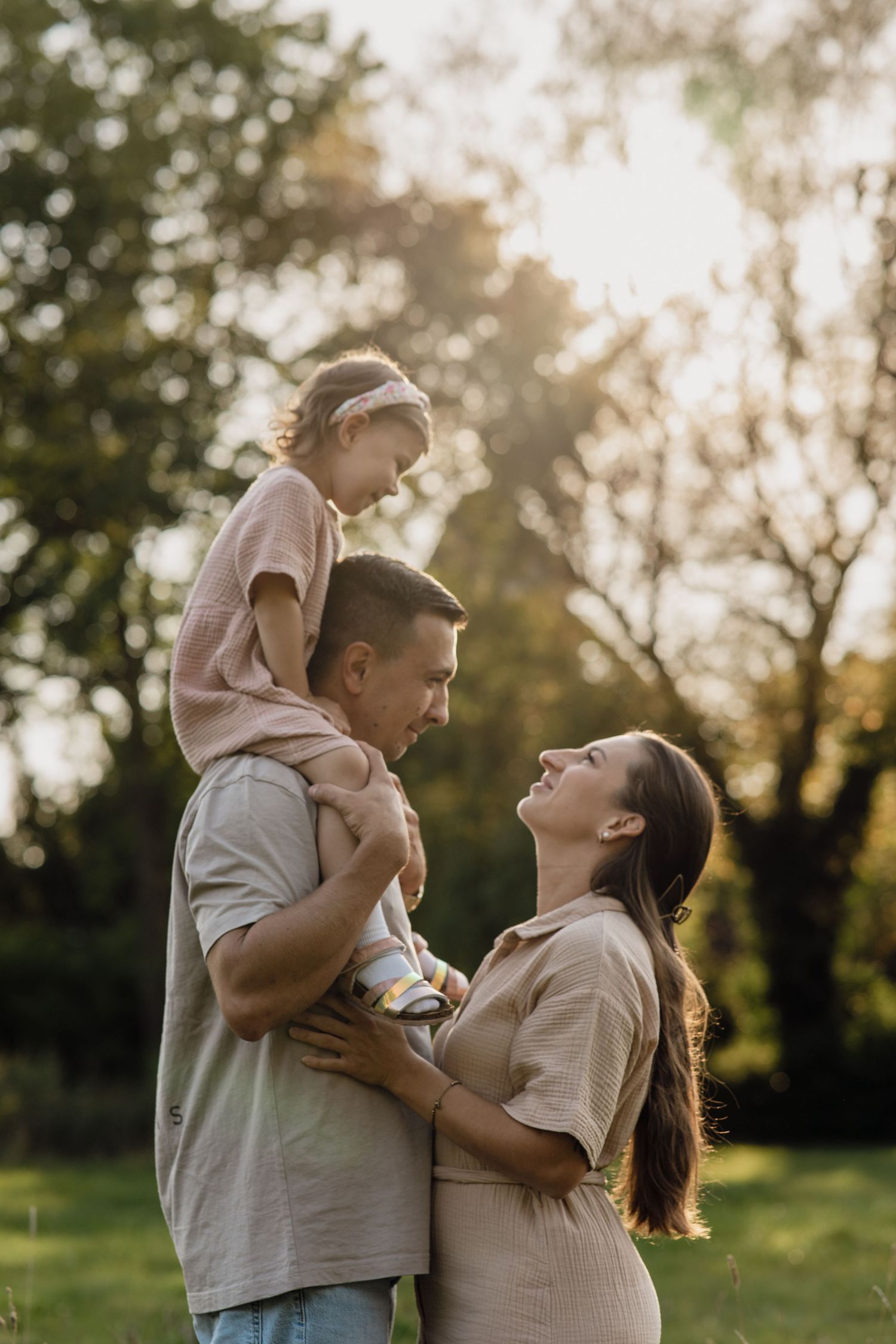 A father with a toddler on his shoulders gazes lovingly at his pregnant wife during a golden hour family photo session.