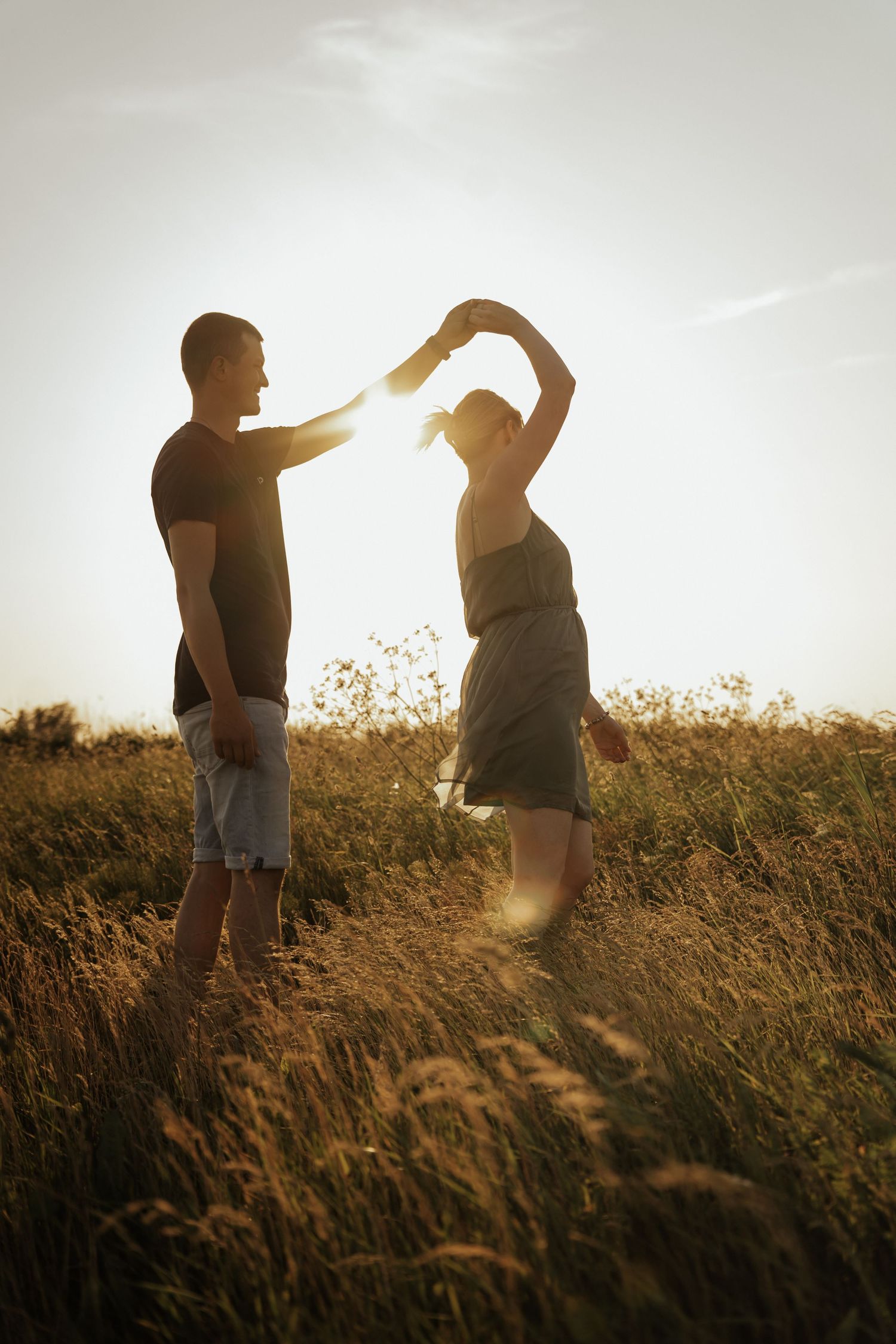 Silhouetted couple dancing in a field of tall grass as the man twirls his partner against a glowing sunset sky.