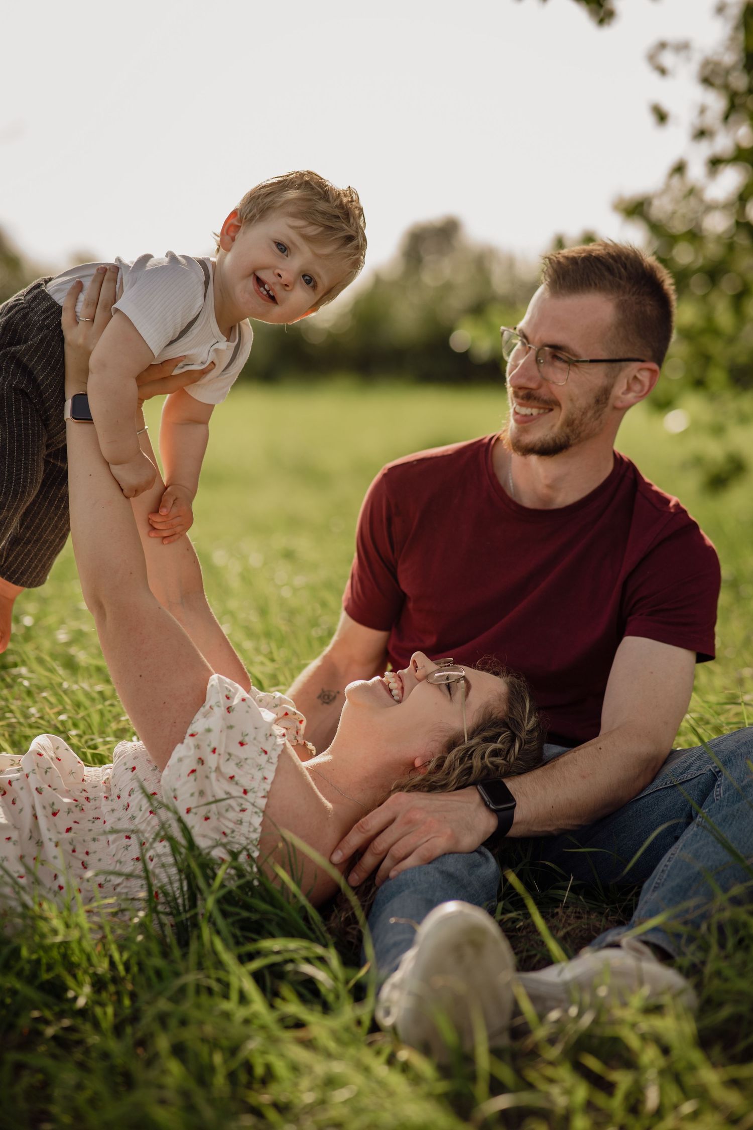 Lächelnde Mutter liegt im Gras und hebt ihr lachendes Kleinkind hoch, während der bärtige Vater daneben in der Sonne sitzt.