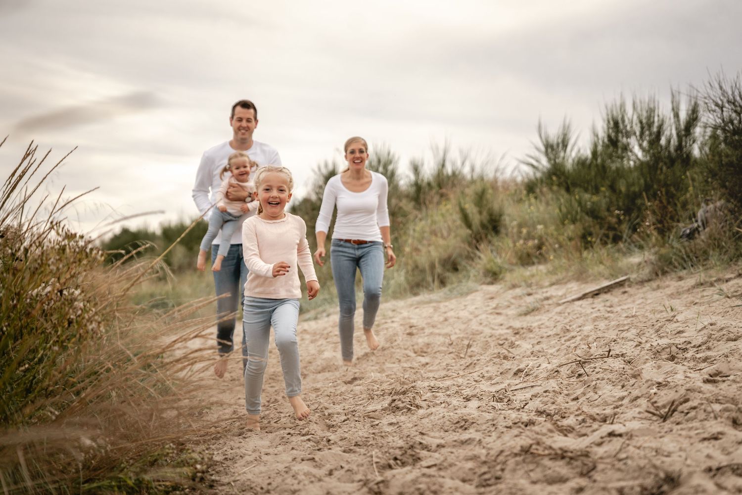 A happy family walks toward the camera along a sandy dune trail, with a girl in front and a father carrying a toddler.