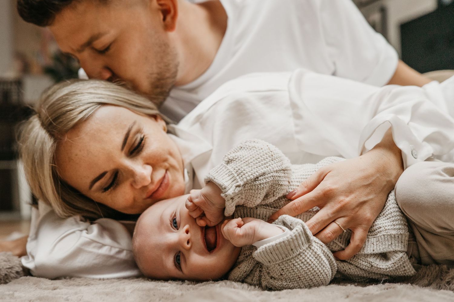 Happy mother and father cuddling and kissing their baby while lying together on a cozy rug in a bright room.