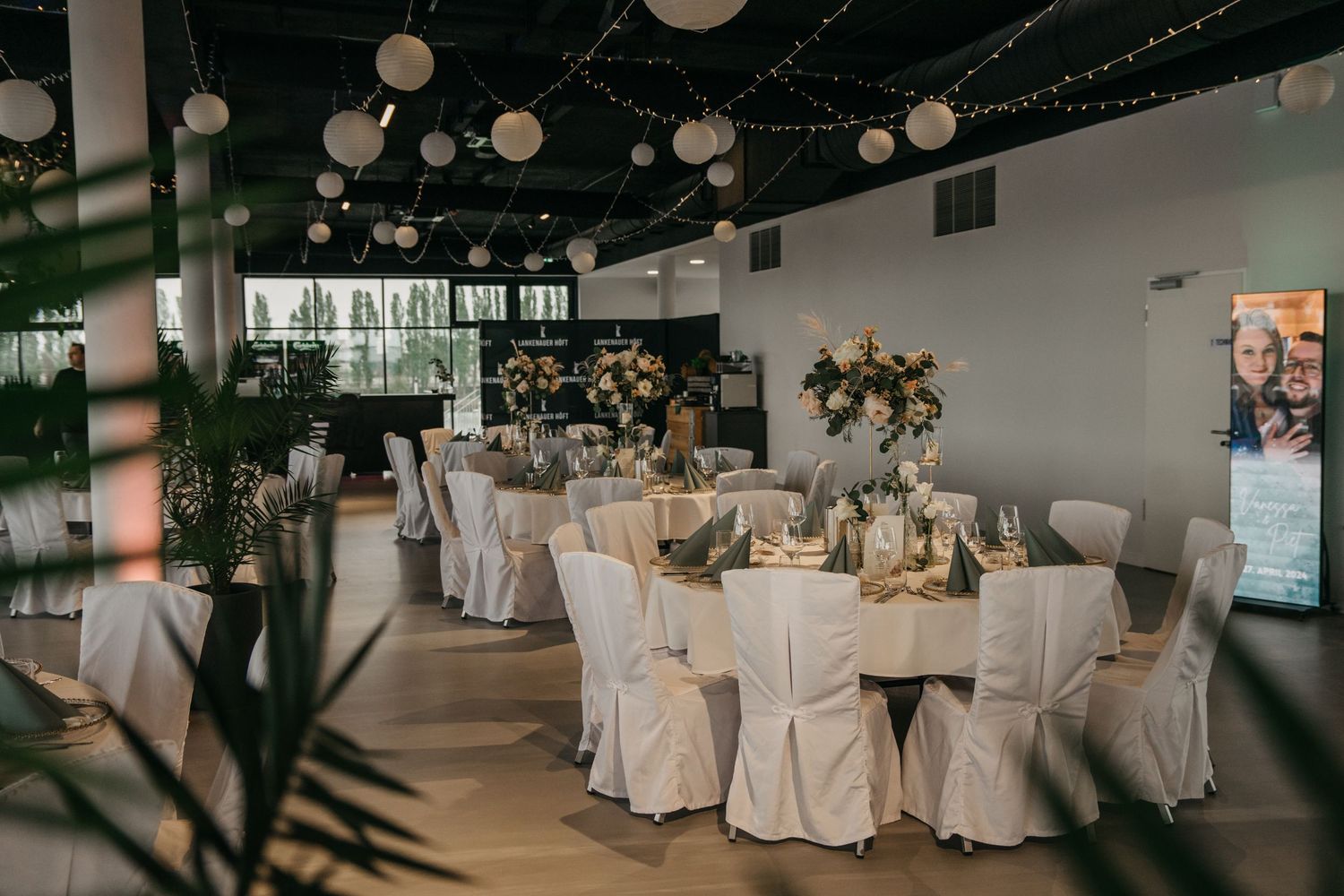 Elegant wedding reception hall with round tables, white chair covers, floral centerpieces and string lights overhead.