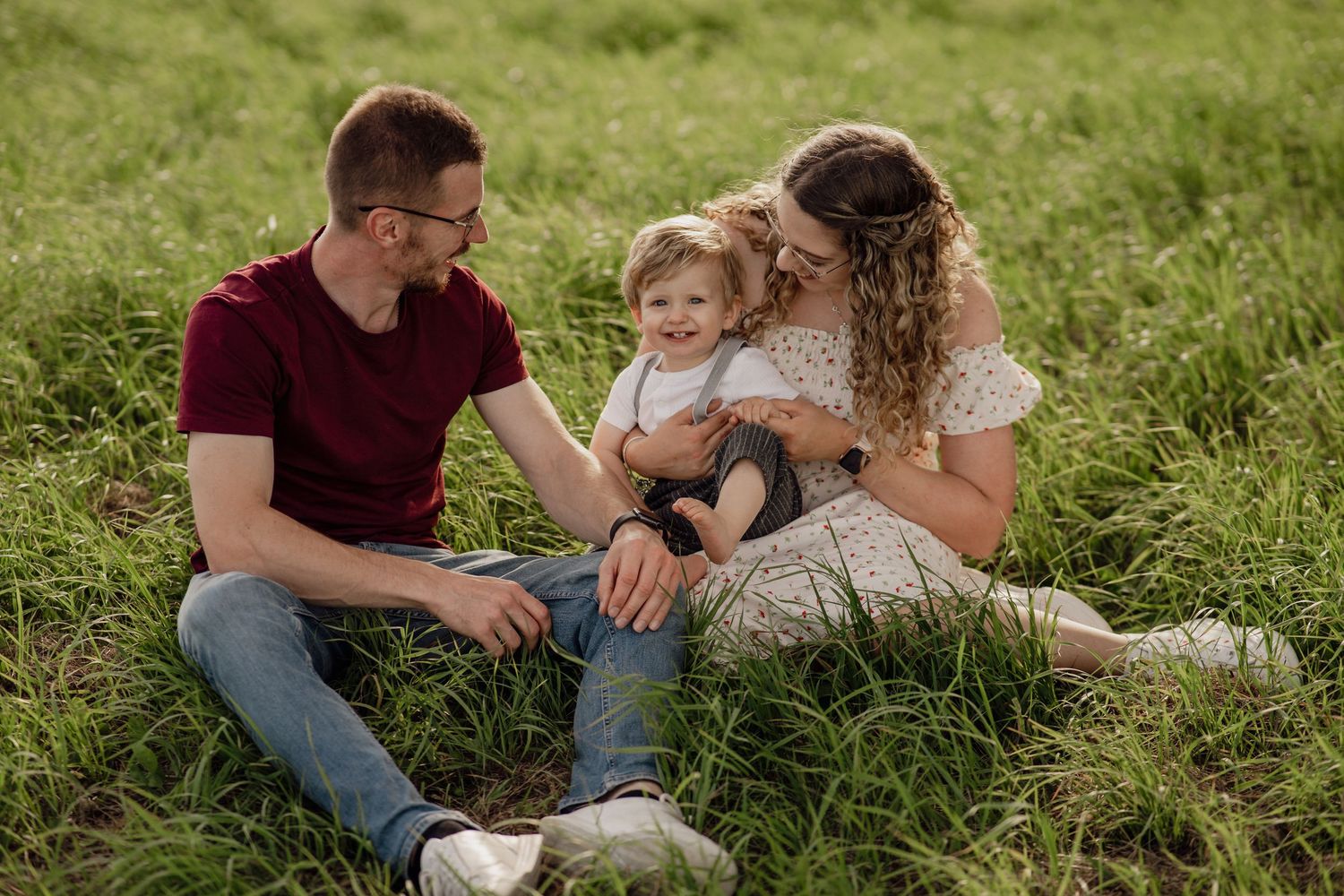 Familie mit Mutter, Vater und kleinem Sohn sitzt zusammen im grünen Gras und lächelt sich liebevoll an.