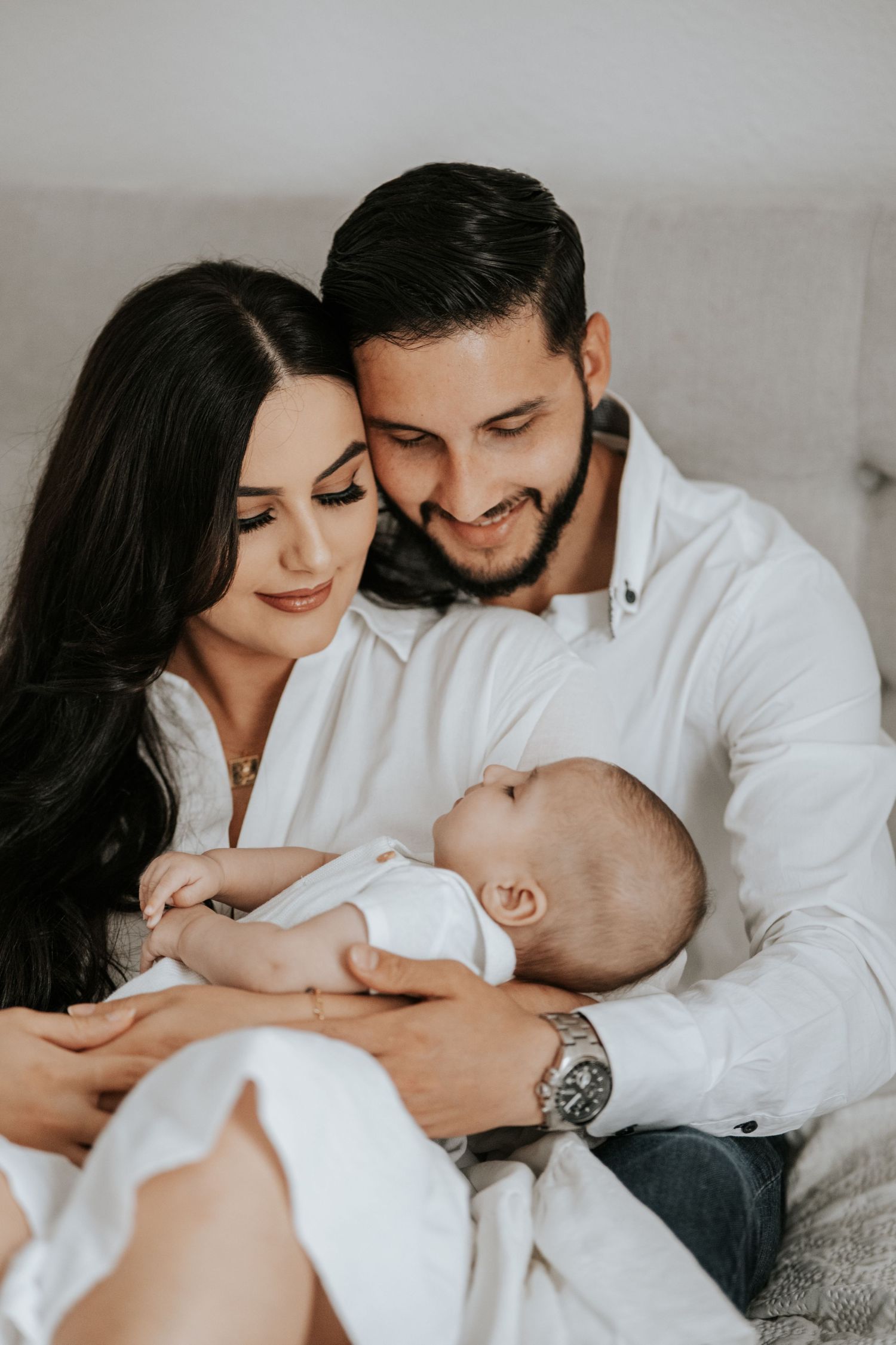 Happy parents in white shirts snuggle close together while holding their sleeping newborn baby.