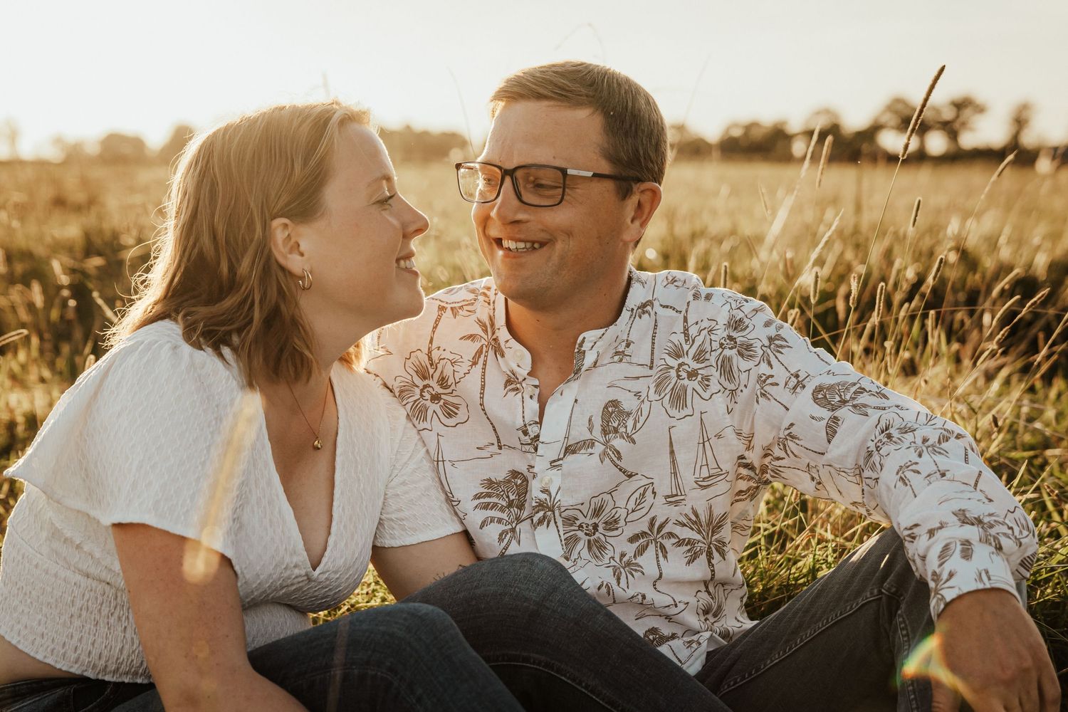 Happy couple laughing and looking at each other while sitting in a sunlit meadow during golden hour.