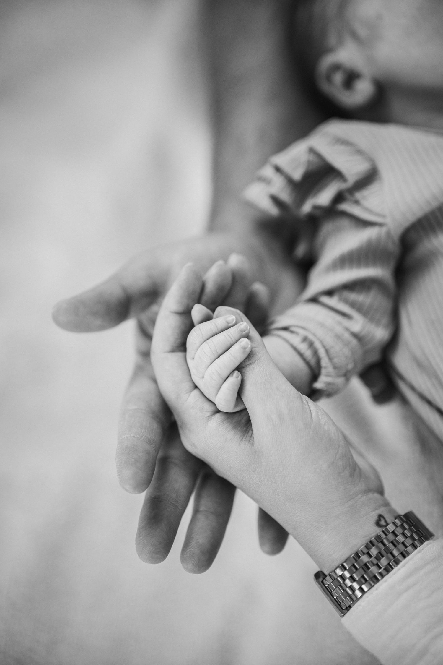Black and white close-up of a parent's hands gently holding a newborn baby's tiny hand, with a watch on the wrist.