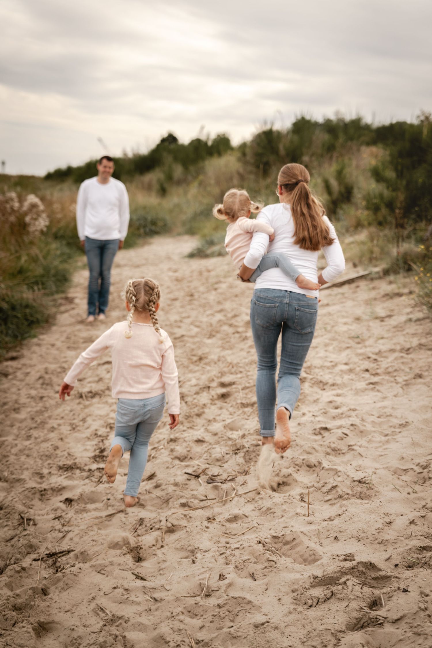 A mother carries a toddler while a young girl runs toward a father waiting on a sandy path through coastal dunes.
