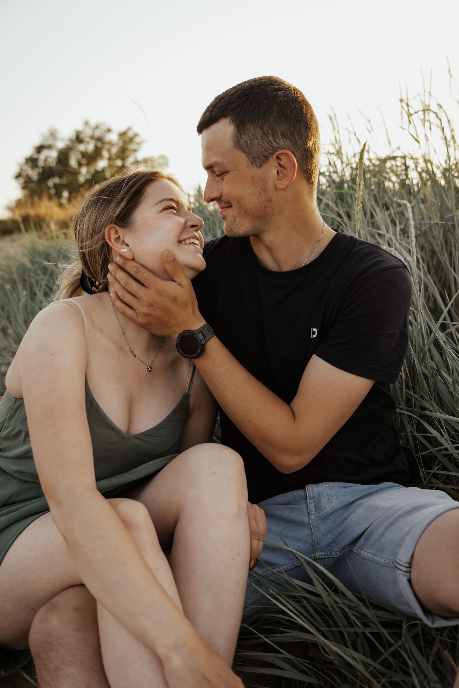Joyful couple sitting among beach grass as the man gently cups the smiling woman's face.