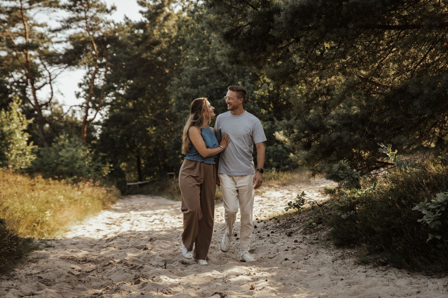 A couple strolls arm in arm along a sandy trail through pine trees and dune grass during an engagement photo session.