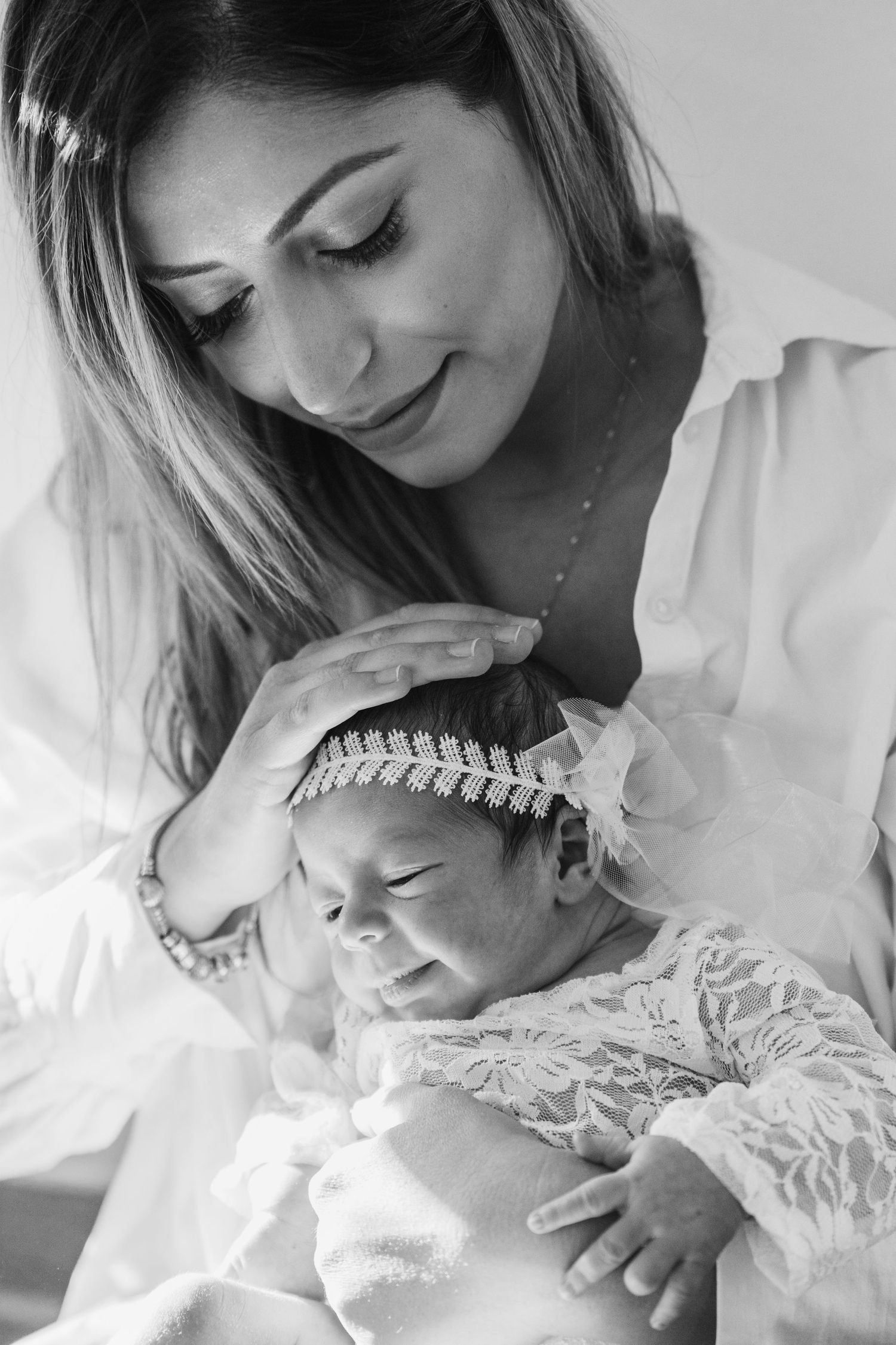 Black and white image of a smiling mother softly touching her sleeping newborn baby's head, wearing a lace outfit and headband.