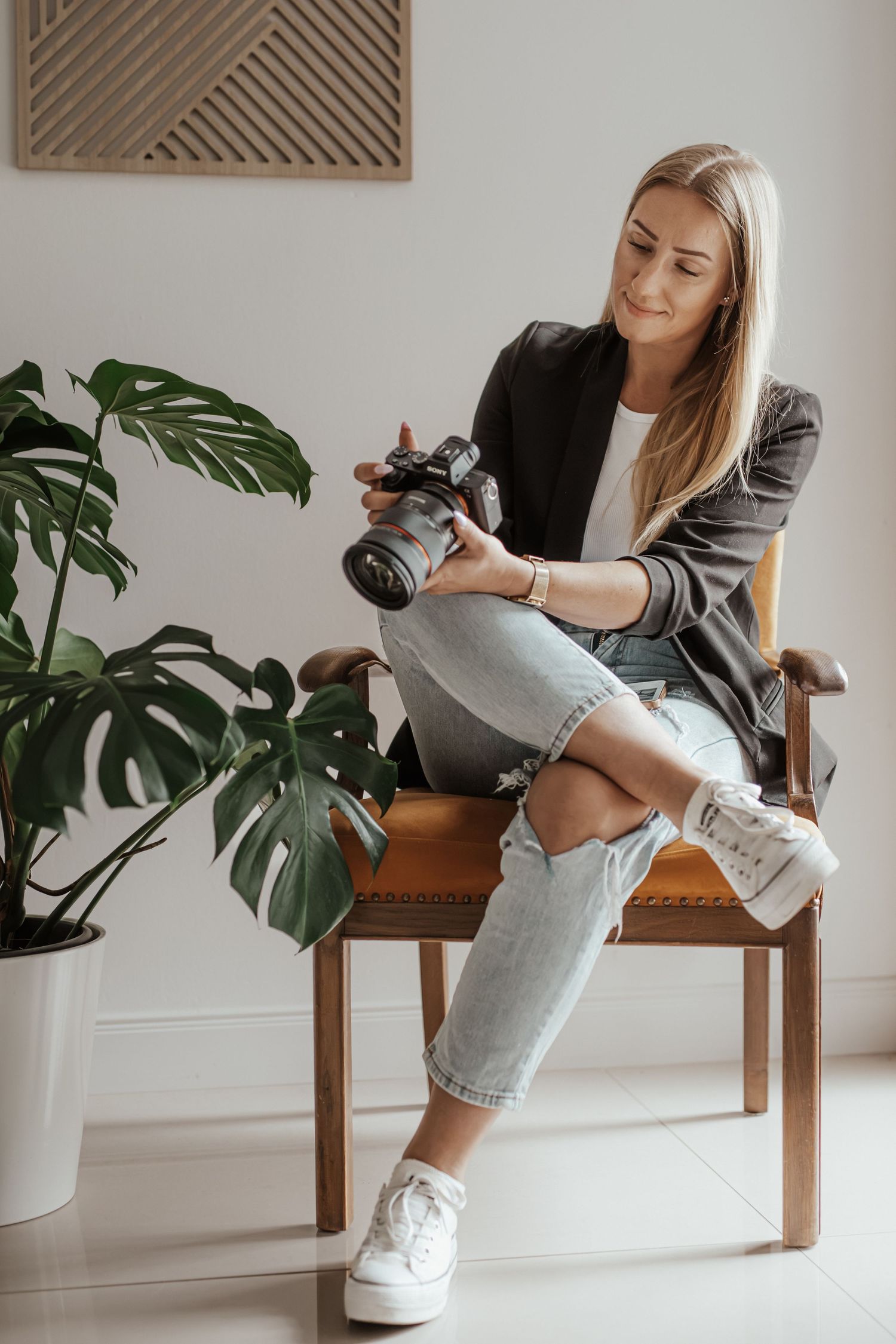 Blonde woman in a blazer and jeans sits on a wooden chair holding a camera, beside a large monstera plant.