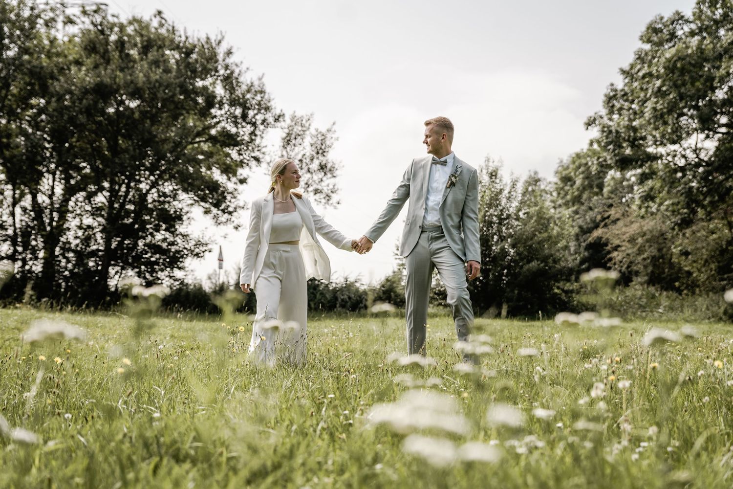 Bride and groom in light outfits hold hands while walking through a green meadow dotted with white flowers on a summer day.