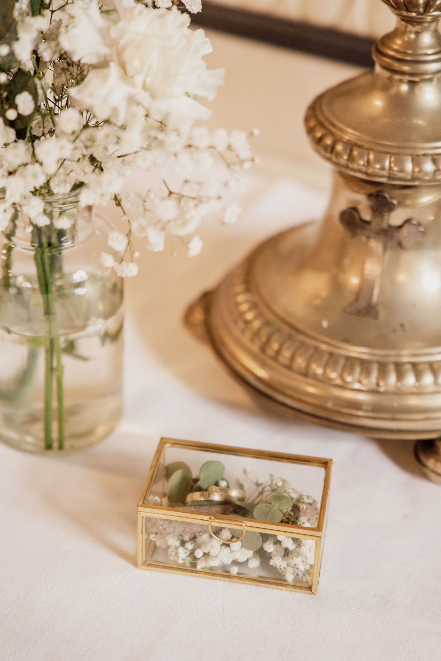 Goldene Ringschachtel mit Eheringen, Eukalyptus und Schleierkraut neben einer Vase mit weißen Blumen und einem Kerzenhalter im Vintage-Stil.