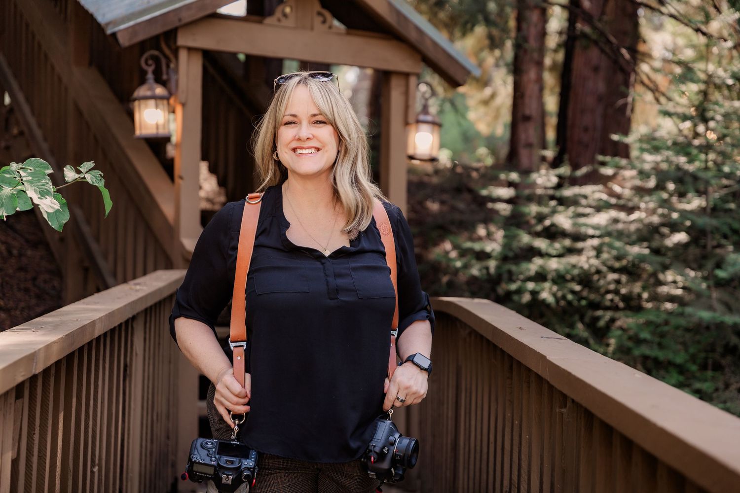 Smiling photographer stands on wooden deck with cameras and forest backdrop.
