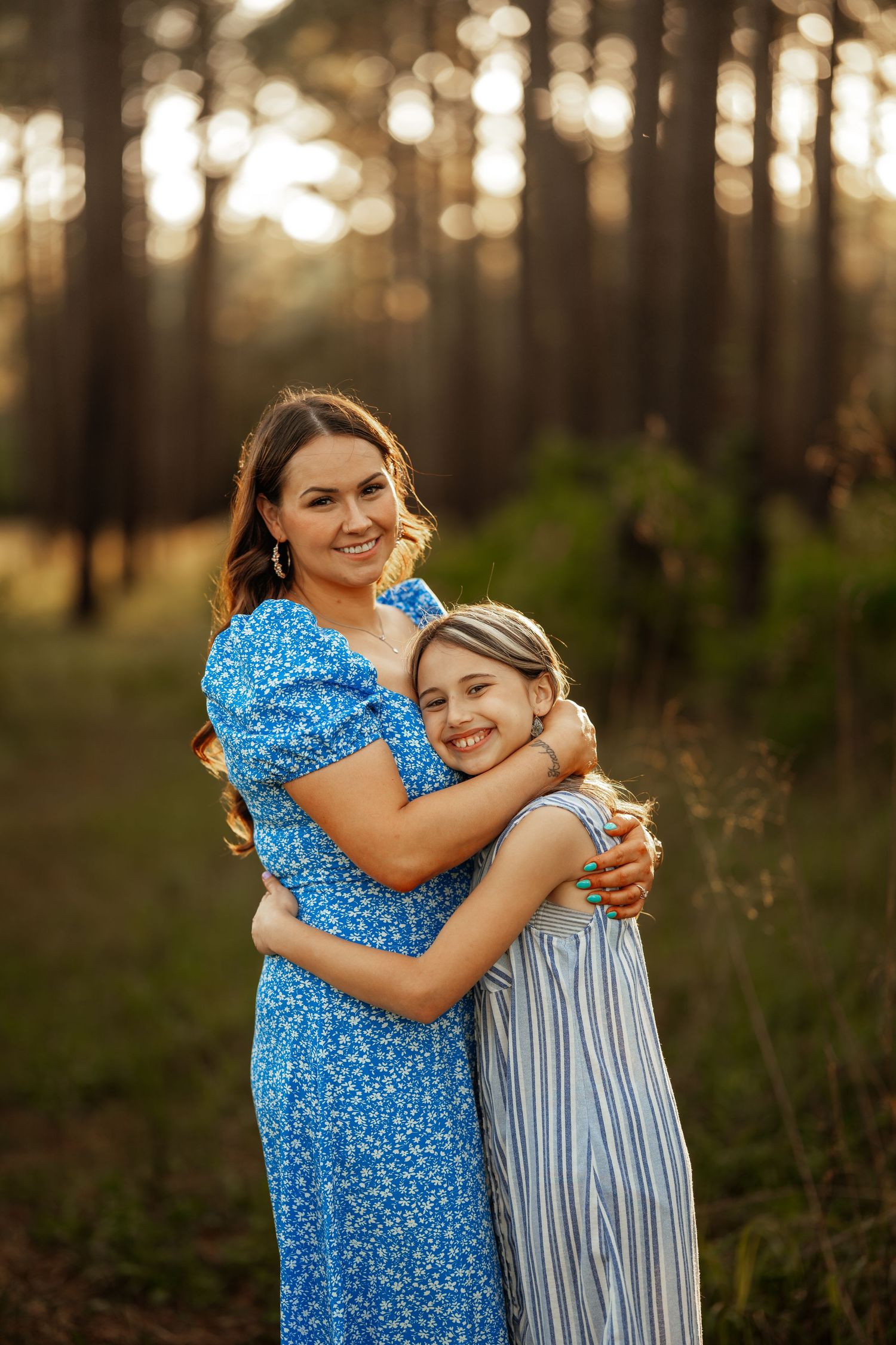 Two people in blue dresses share a warm embrace while posing for a portrait in a forest at sunset.