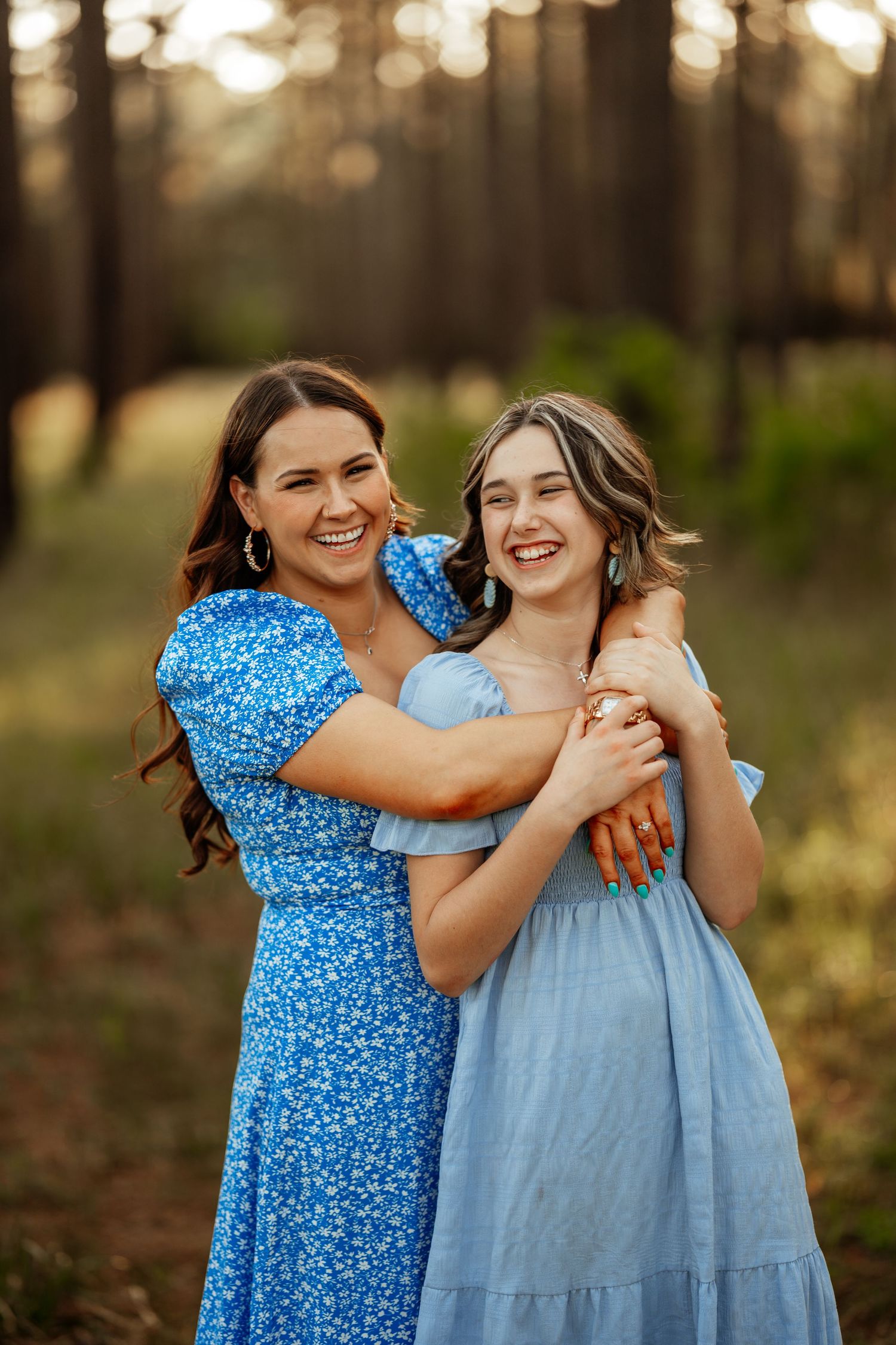 Two people wearing coordinating blue dresses embrace while laughing together in a wooded setting.