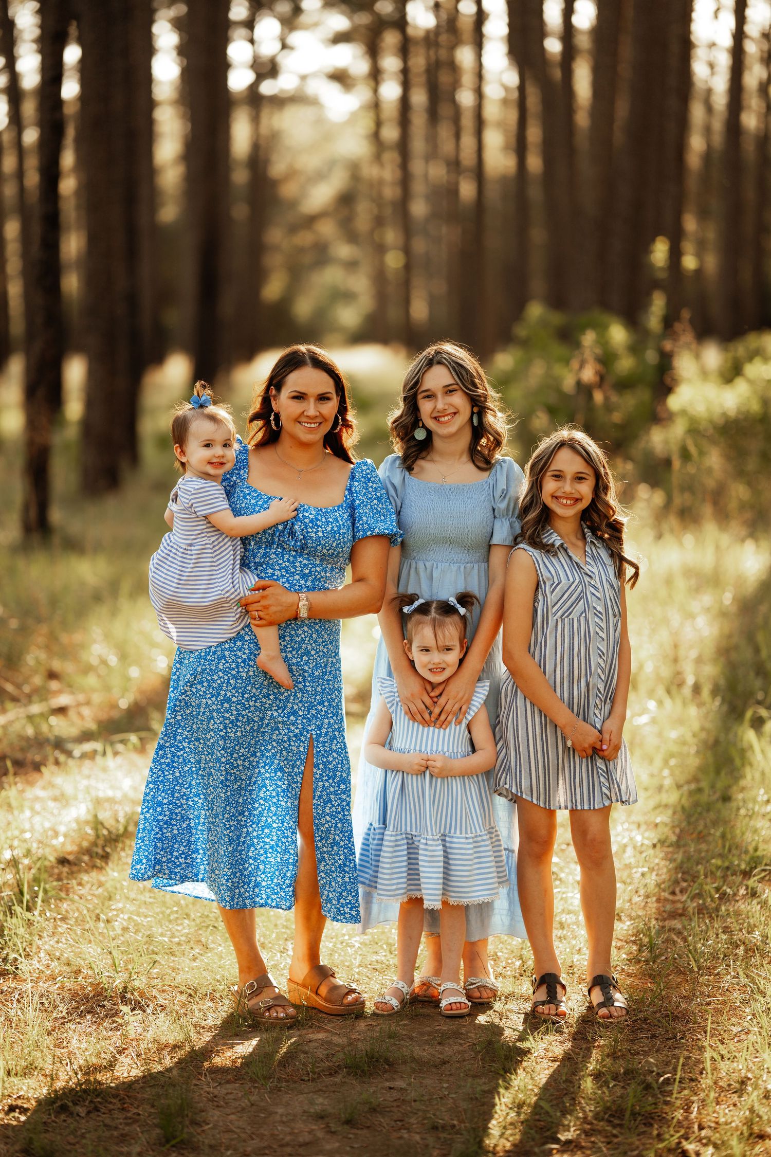 Five people wearing coordinating blue and white dresses pose together on a forest path at sunset.