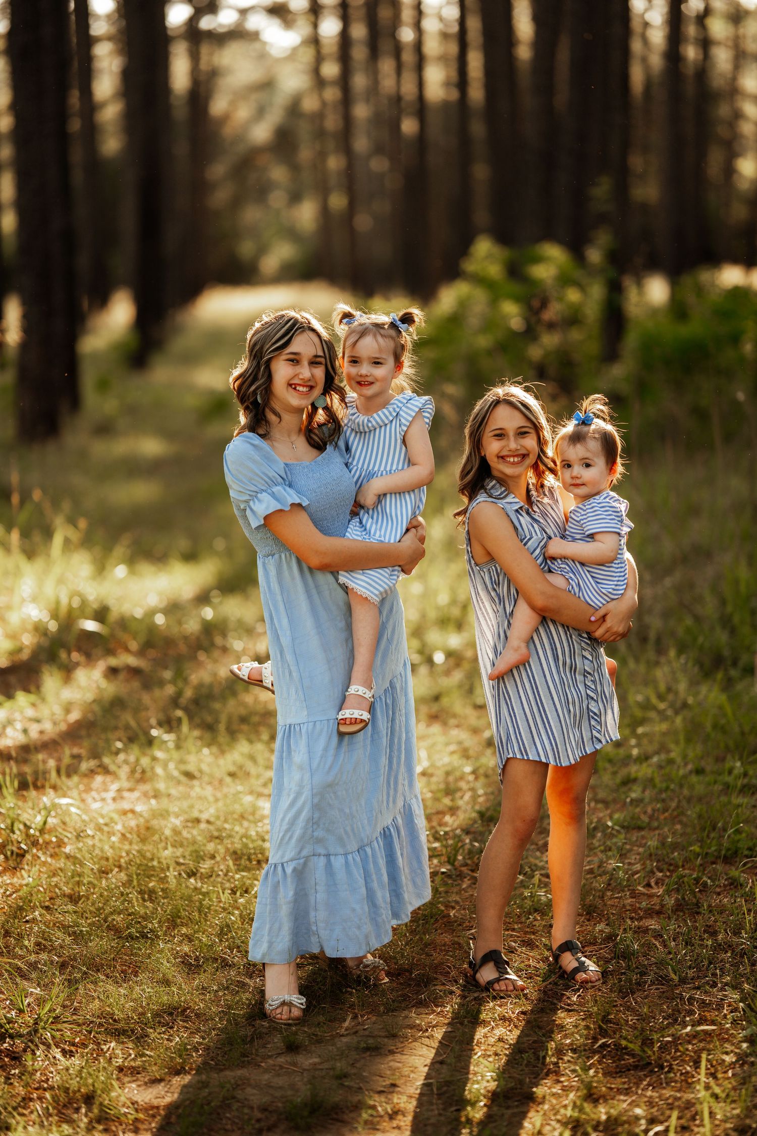 Two figures in blue dresses holding small children pose together on a forest path at sunset.