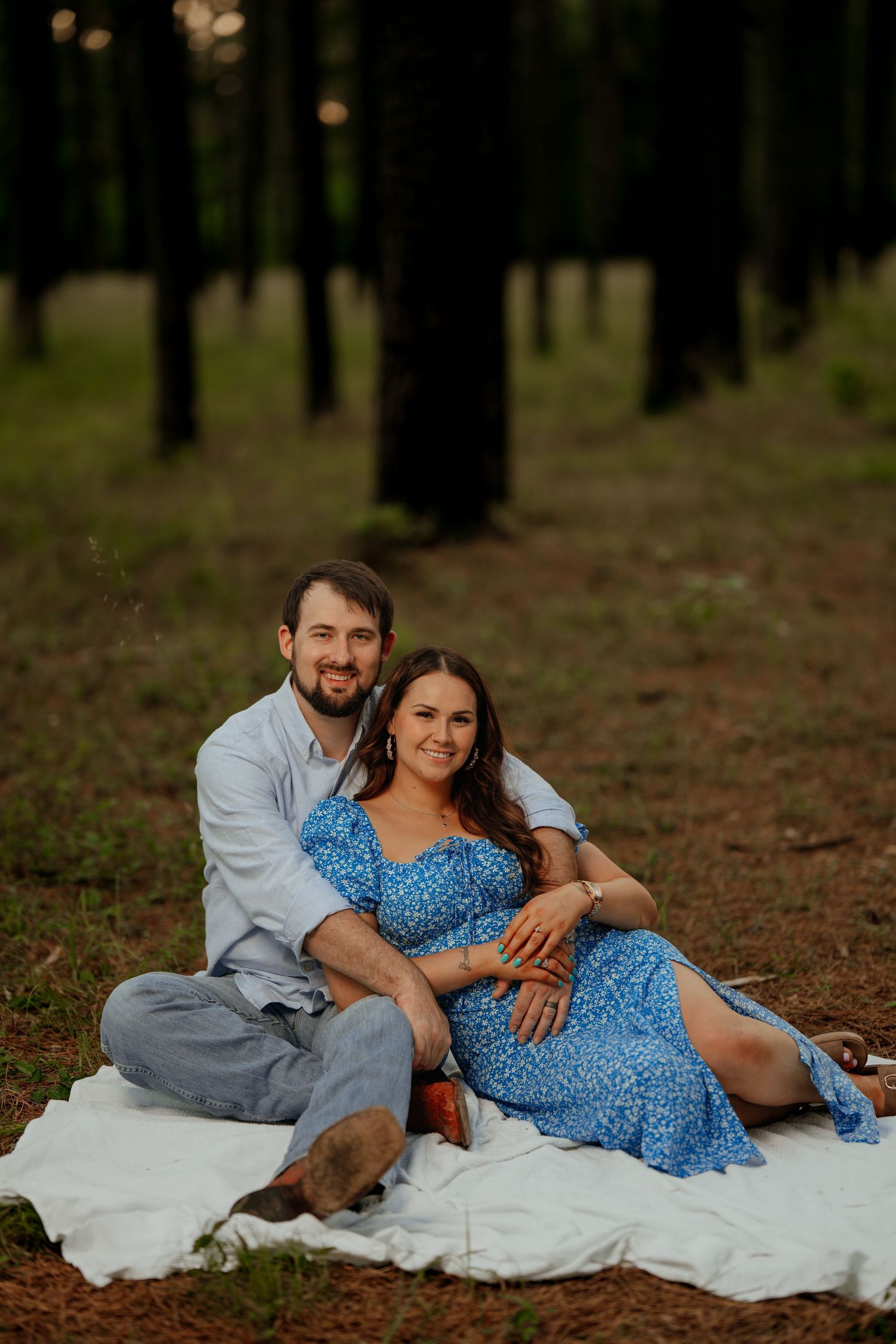 A couple sits together on a white blanket in a serene pine forest setting during a romantic photoshoot.