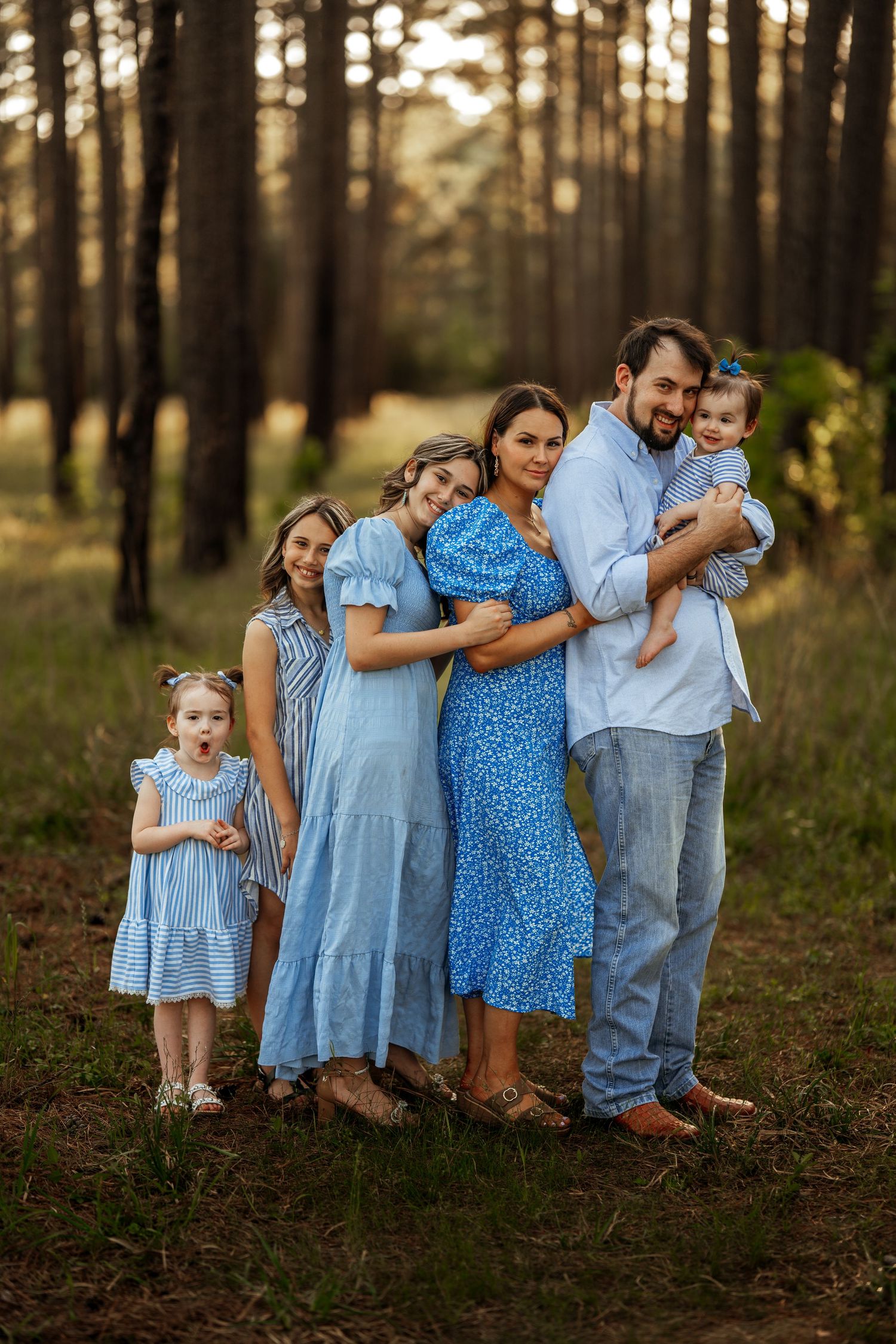 A family wearing coordinating blue outfits stands together in a wooded area at sunset.