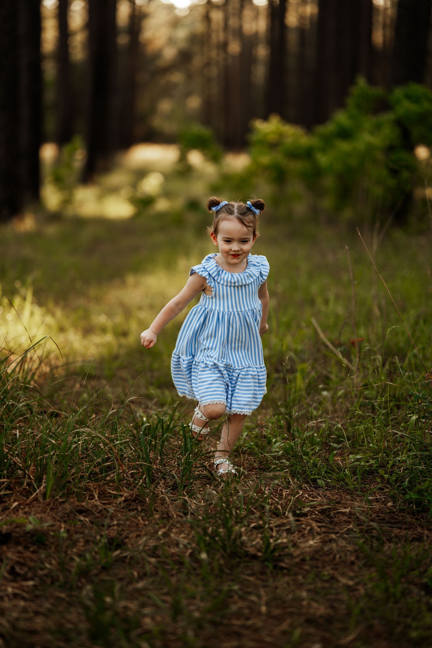 A small child in a blue striped dress runs playfully through a sunlit forest path.