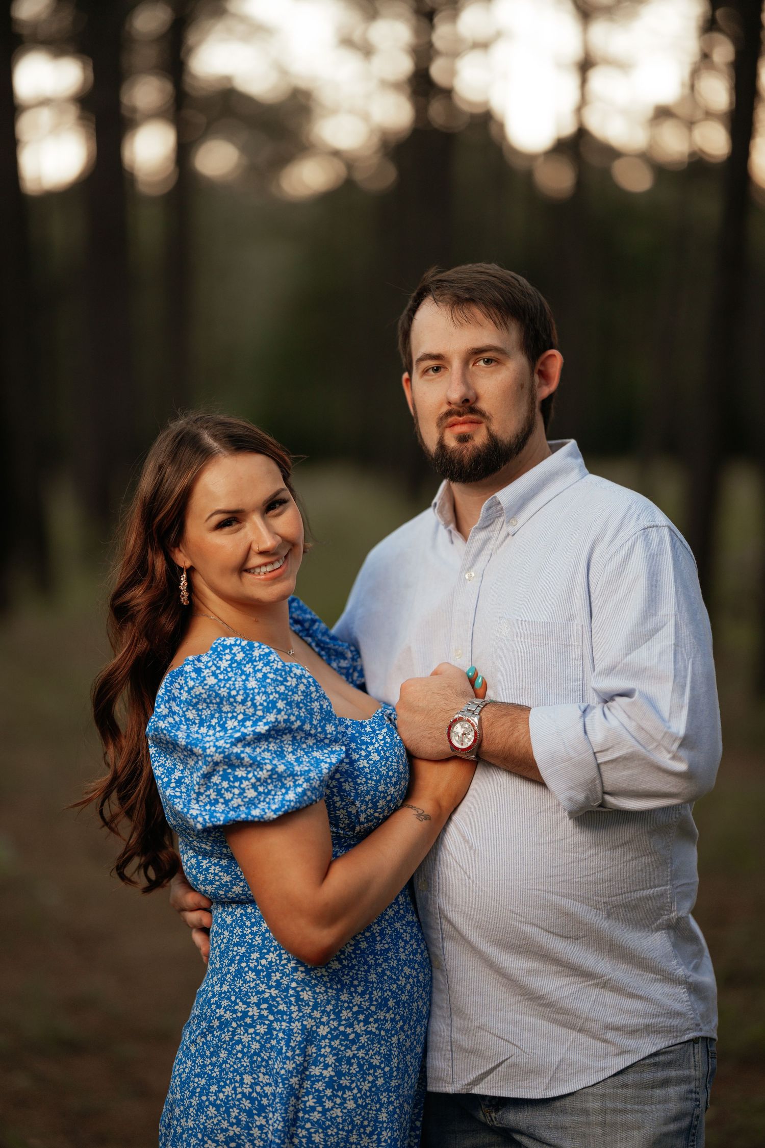 A couple poses together in a wooded setting at sunset, wearing a blue floral dress and light button-down shirt.