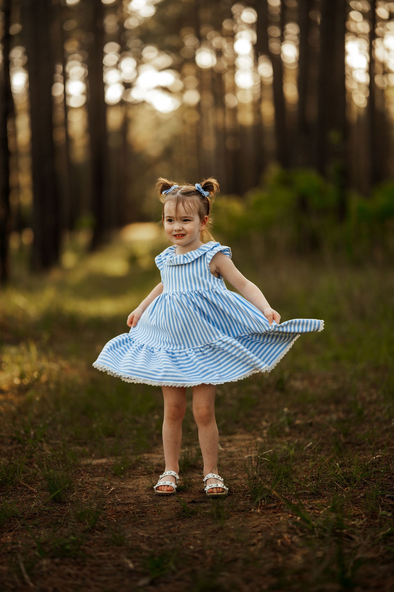 A small child in a light blue striped dress twirls joyfully on a forest path during golden hour.