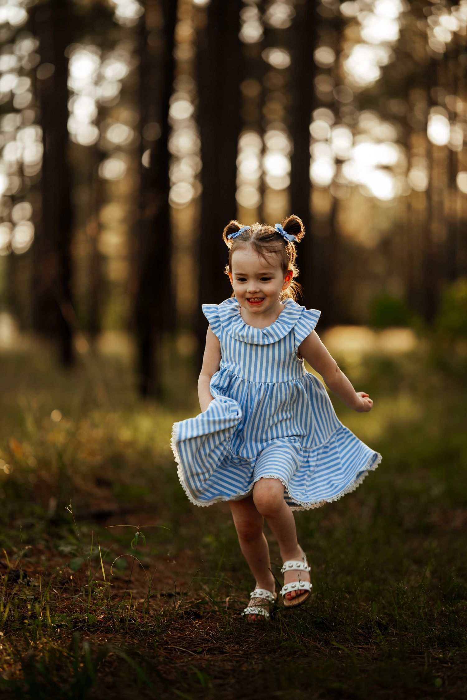 Young child in striped blue dress twirls playfully in a sunlit forest setting.
