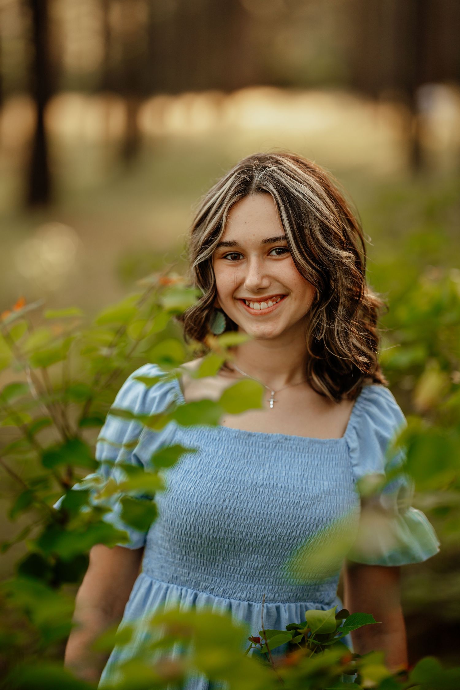 Young woman in blue smocked dress poses outdoors among green foliage in natural lighting.
