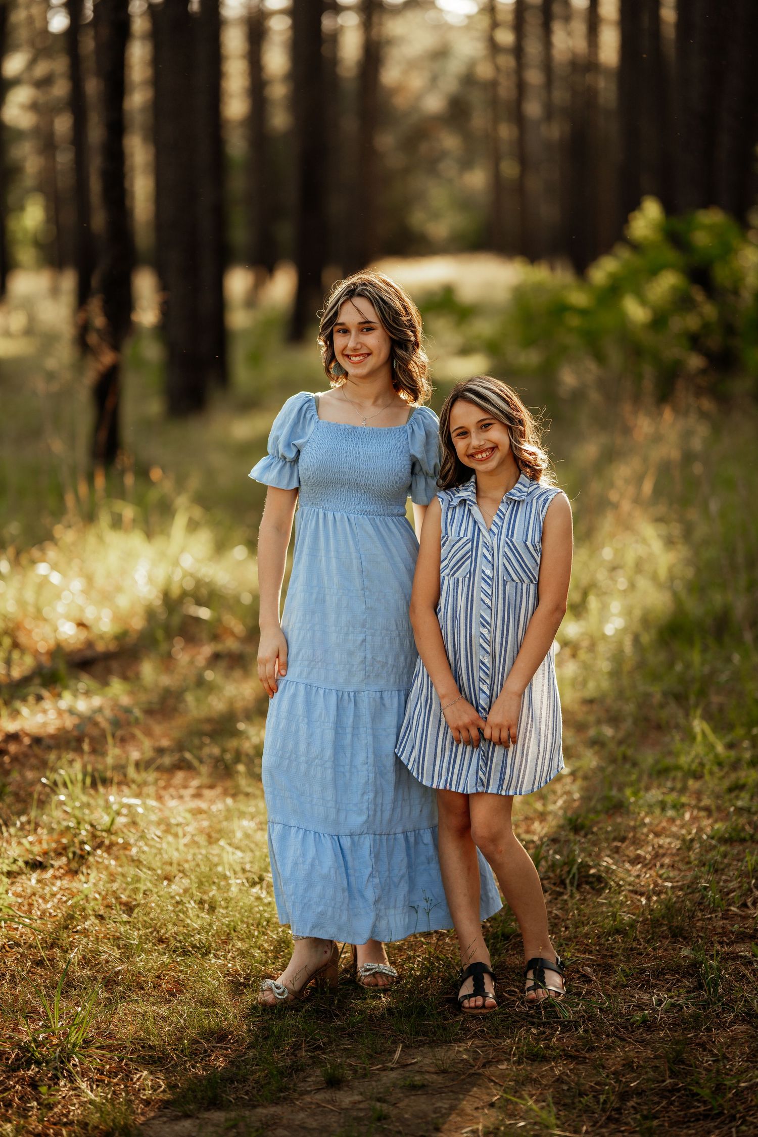 Two figures pose together in a sunlit forest wearing blue and white dresses against a backdrop of tall pine trees.