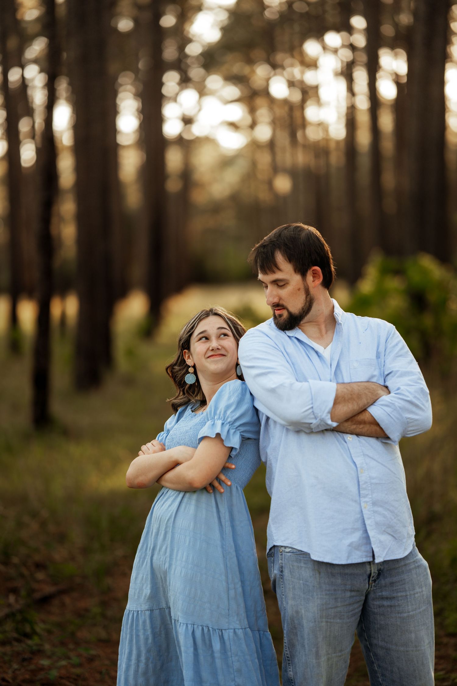 A couple in casual attire poses playfully with crossed arms in a sunlit forest.