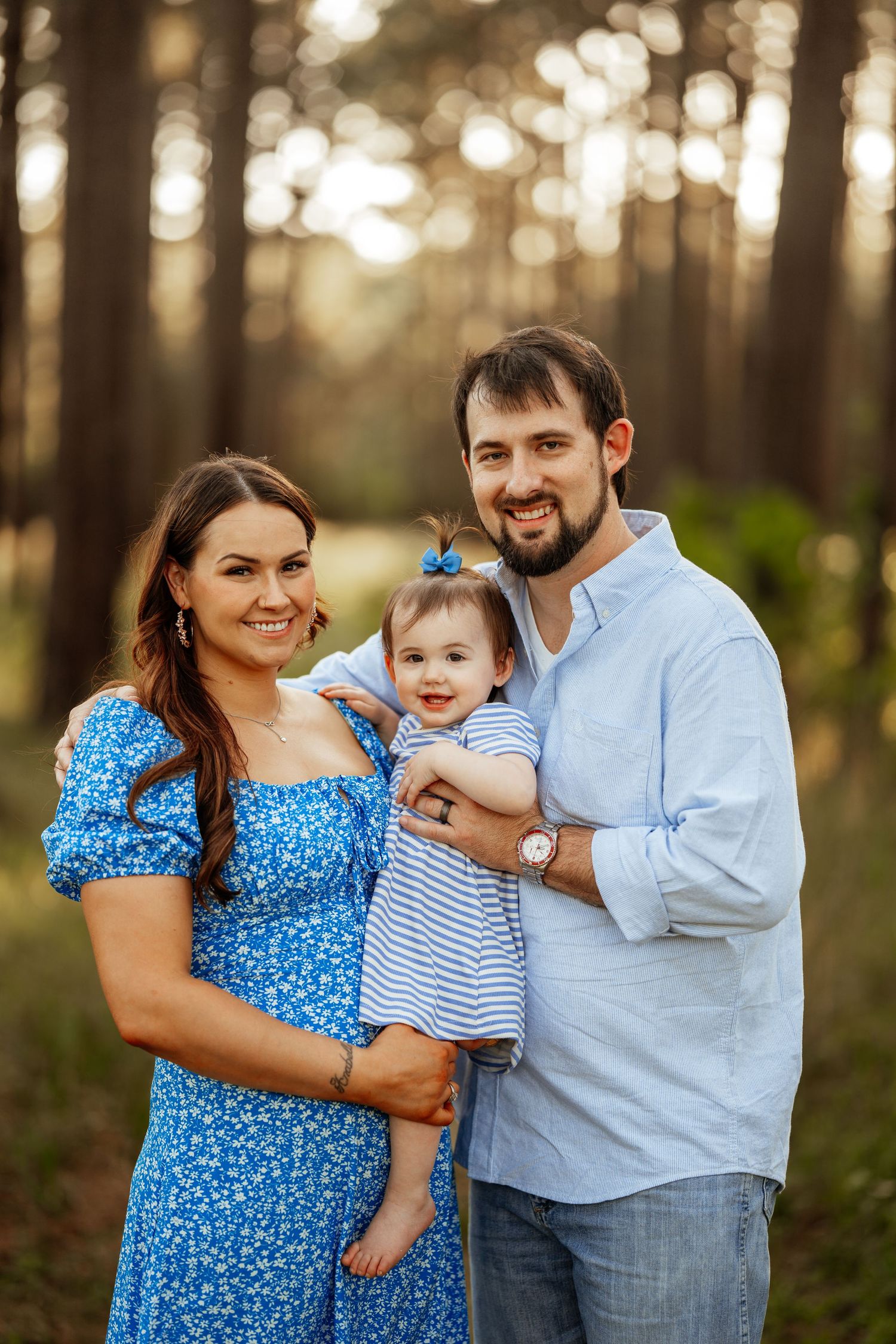 A family in coordinating blue outfits poses together in a sunlit forest setting during golden hour.