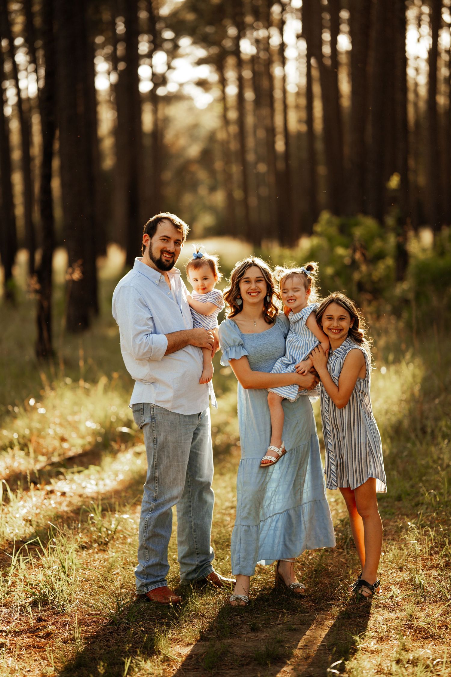 Family of five poses together in a sunlit forest wearing coordinated blue and white outfits.