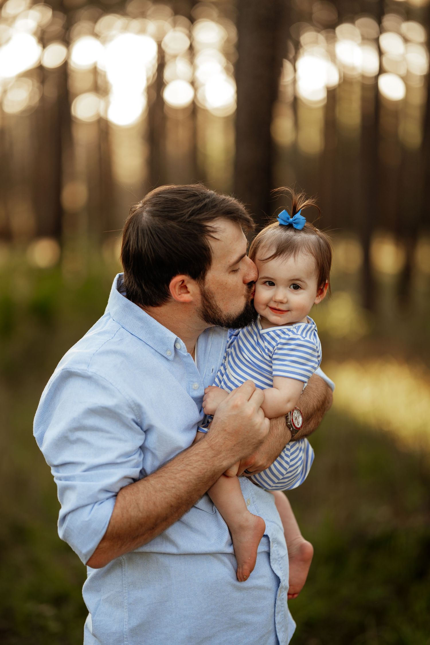 A tender moment between an adult and small child in matching blue outfits captured in a sunlit forest setting.
