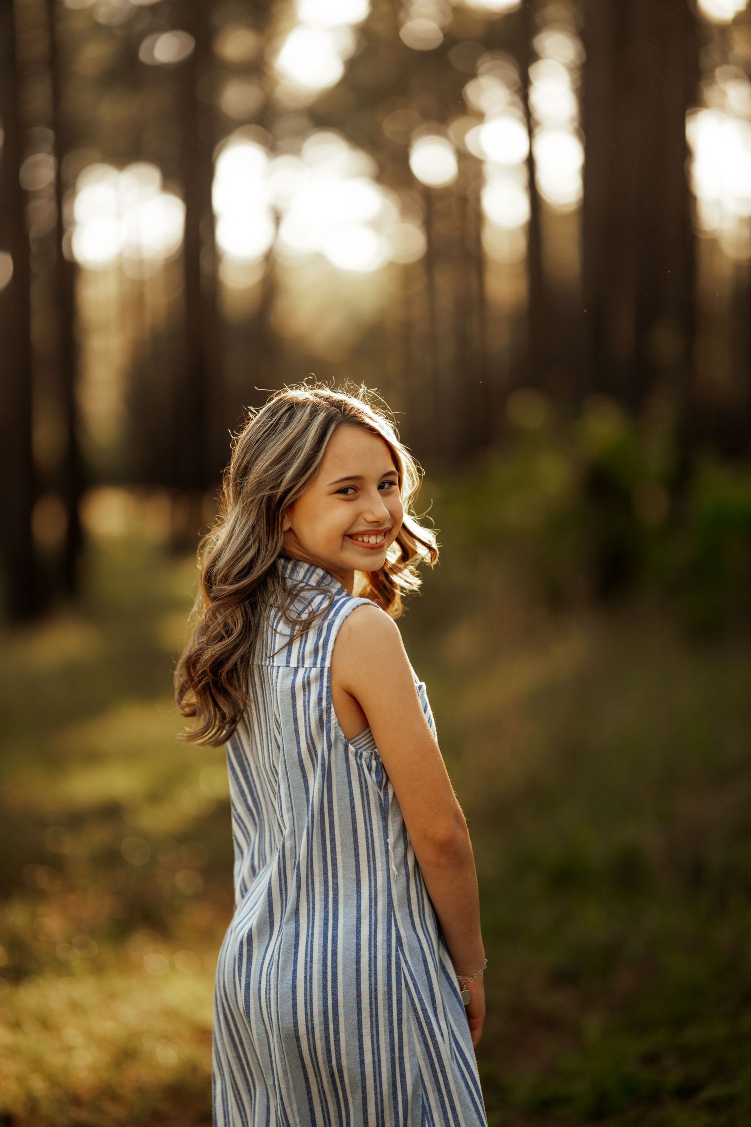 Young person in a striped summer dress poses in a sunlit forest during golden hour.