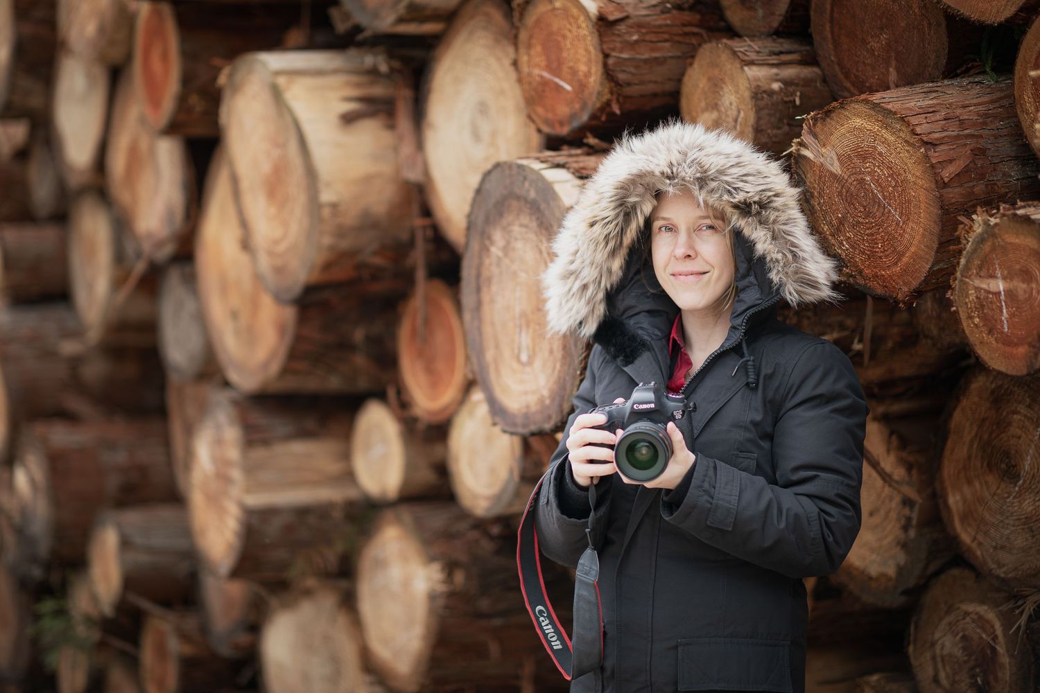 Photographer in winter coat with fur hood stands in front of stacked wood logs.
