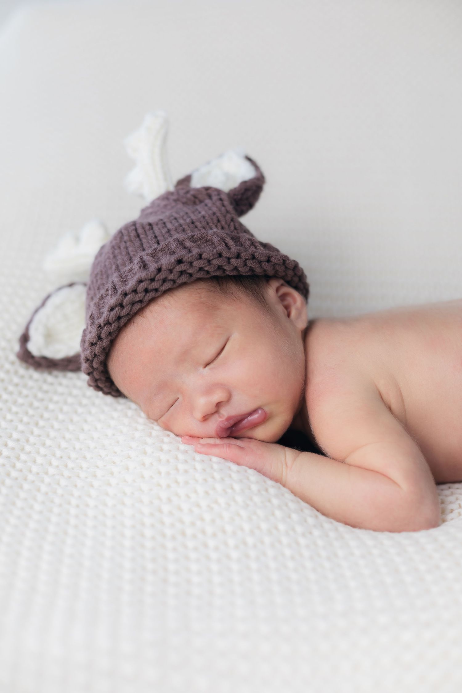 A sleeping newborn wearing a knitted brown animal hat rests on white textured bedding.