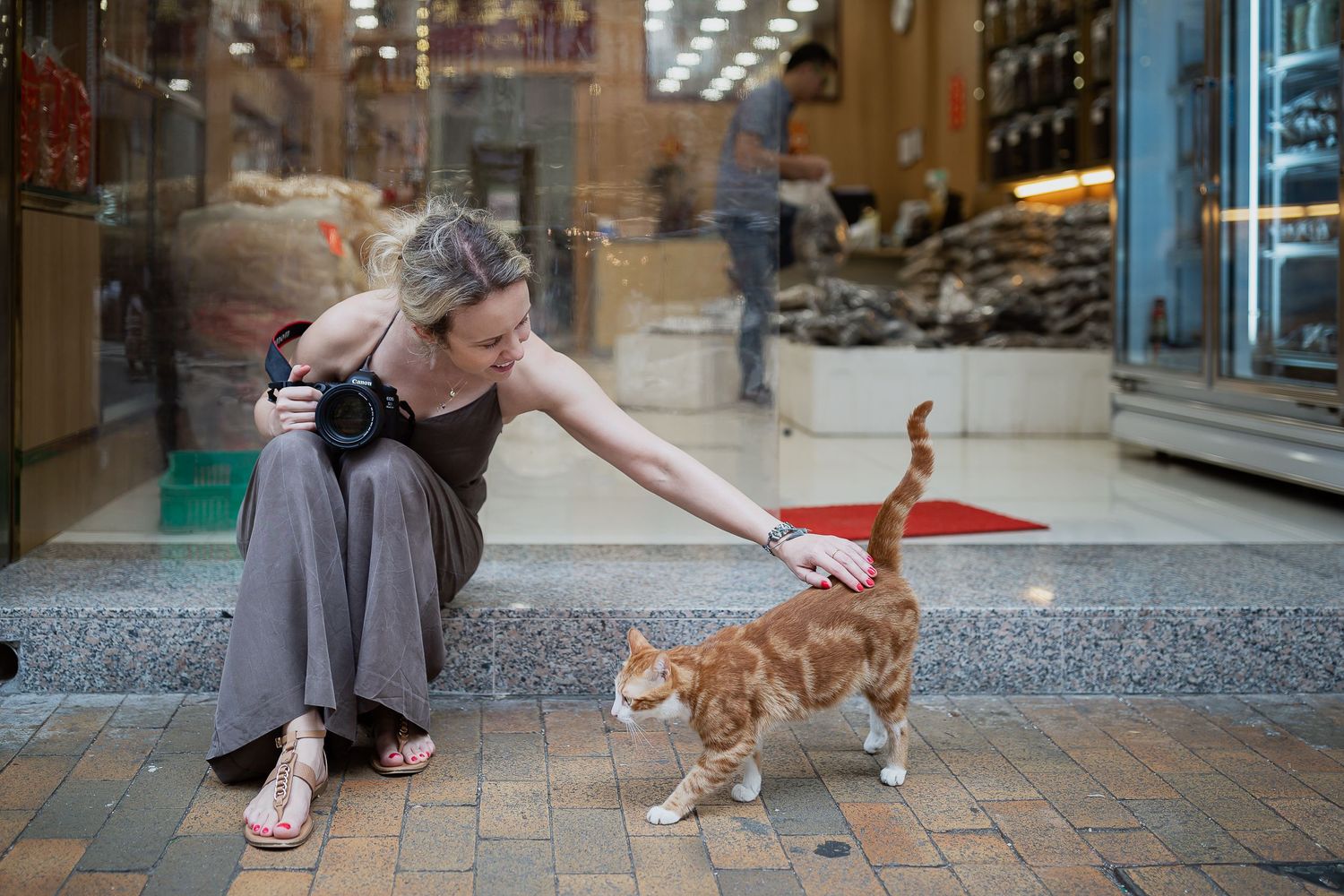Person in gray dress reaches out to pet orange cat on urban sidewalk.