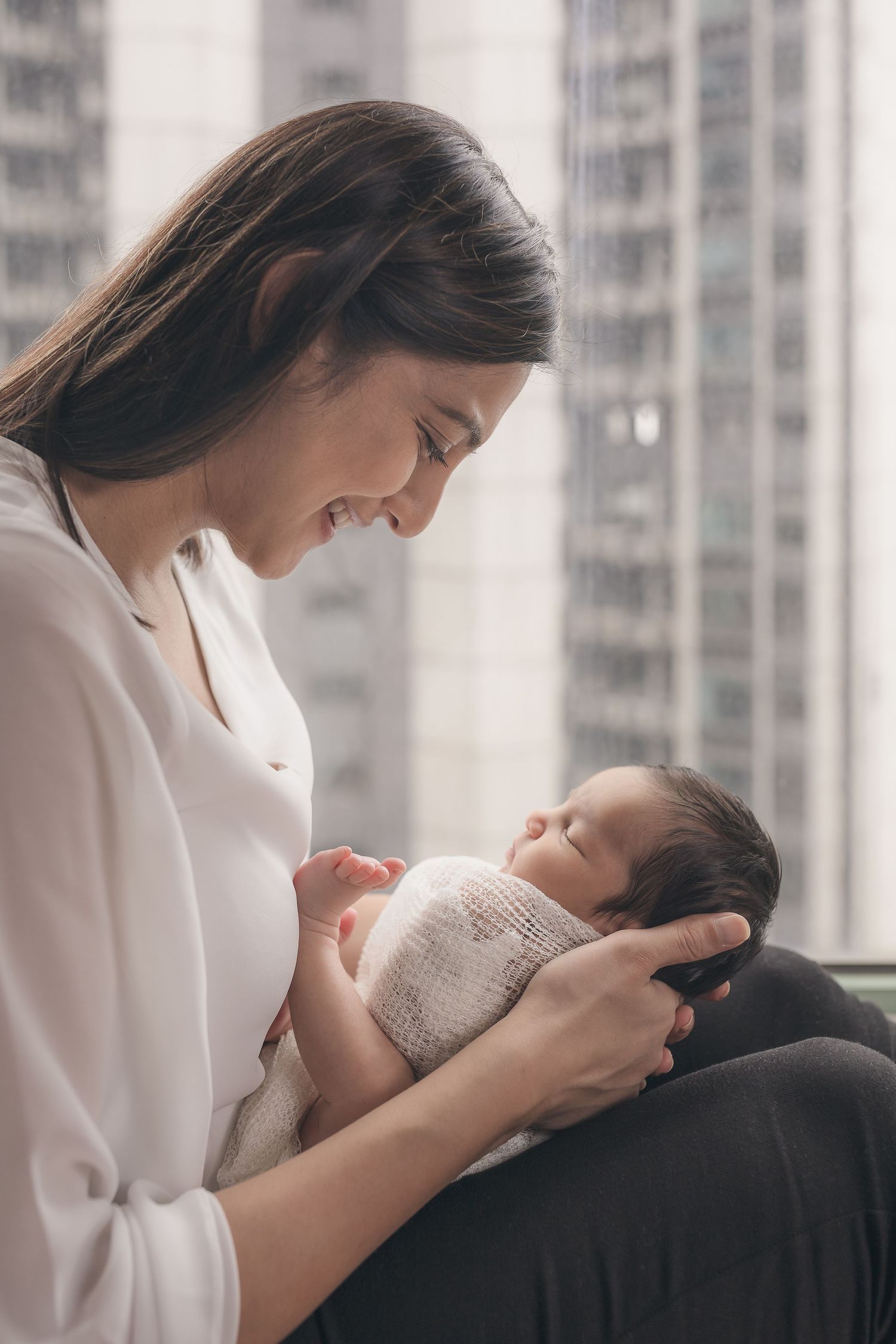 A tender moment shows a parent cradling a sleeping newborn by a window with city views.