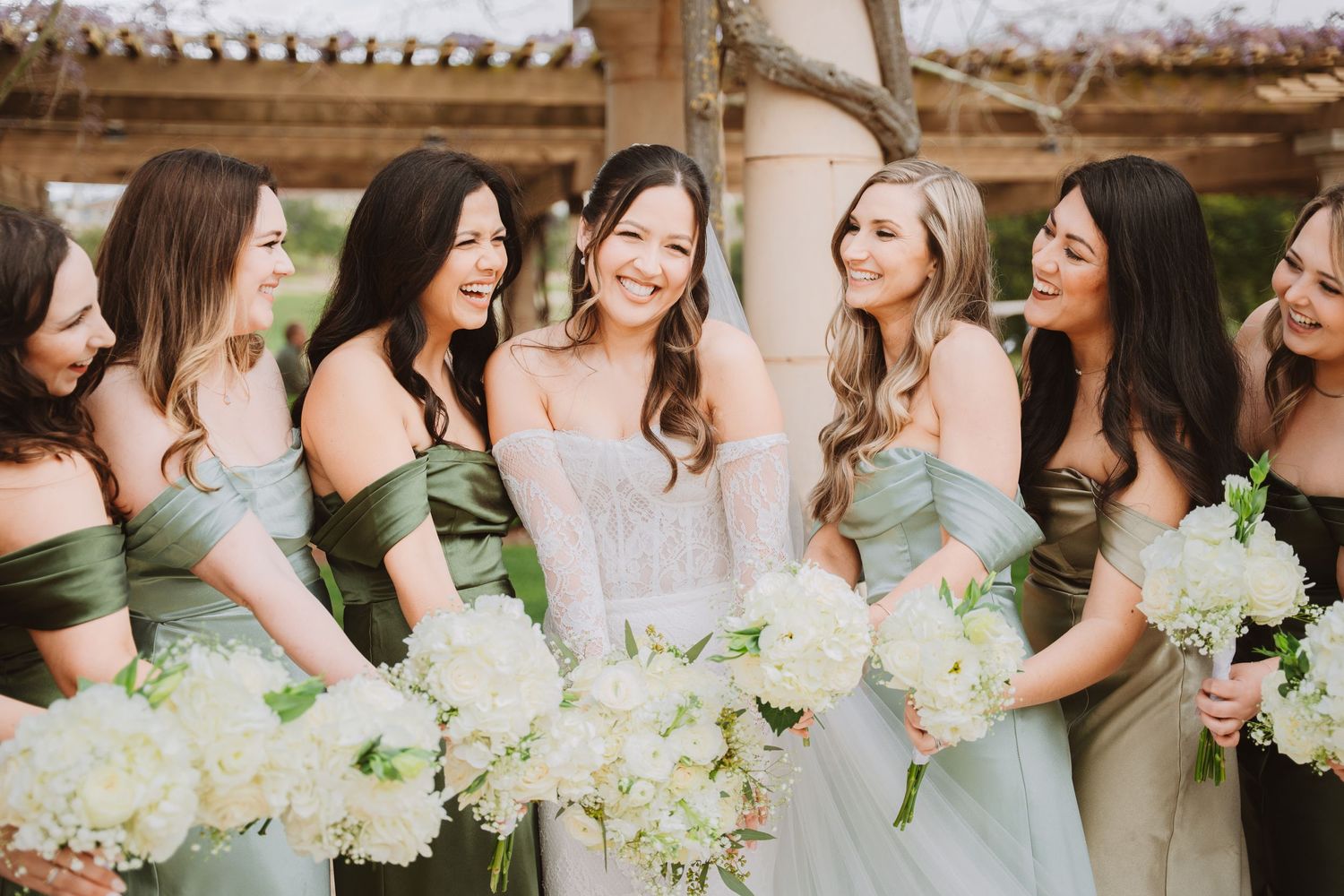 A wedding party in sage green dresses holds white hydrangea bouquets while sharing a joyful moment together.