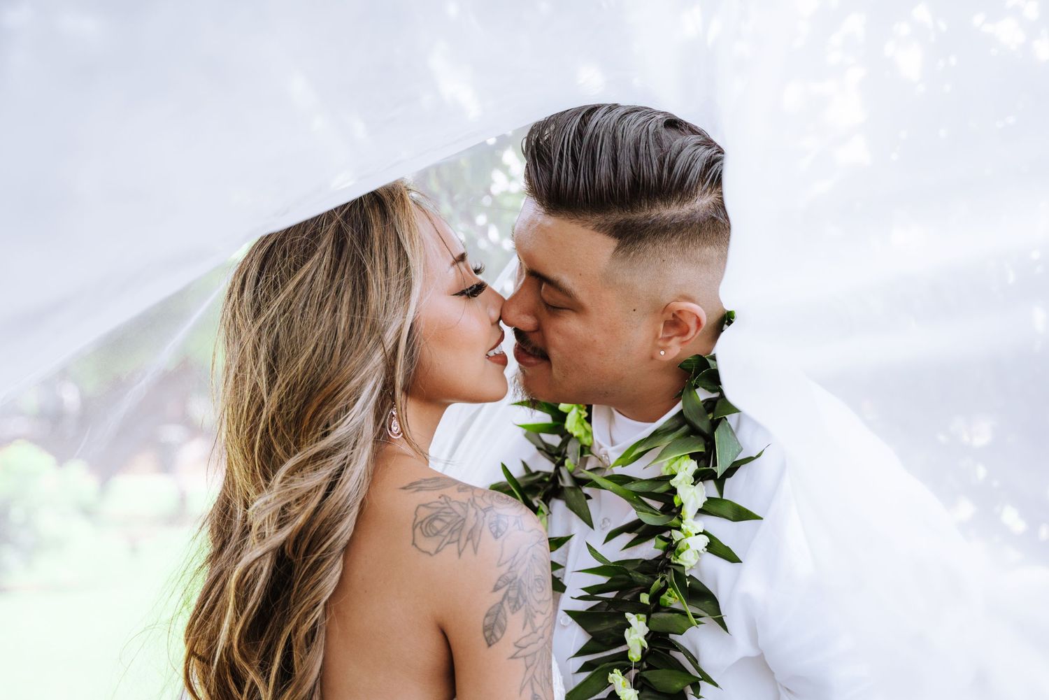 A romantic Hawaiian wedding couple sharing a tender moment with traditional leaf leis against a soft dreamy background.
