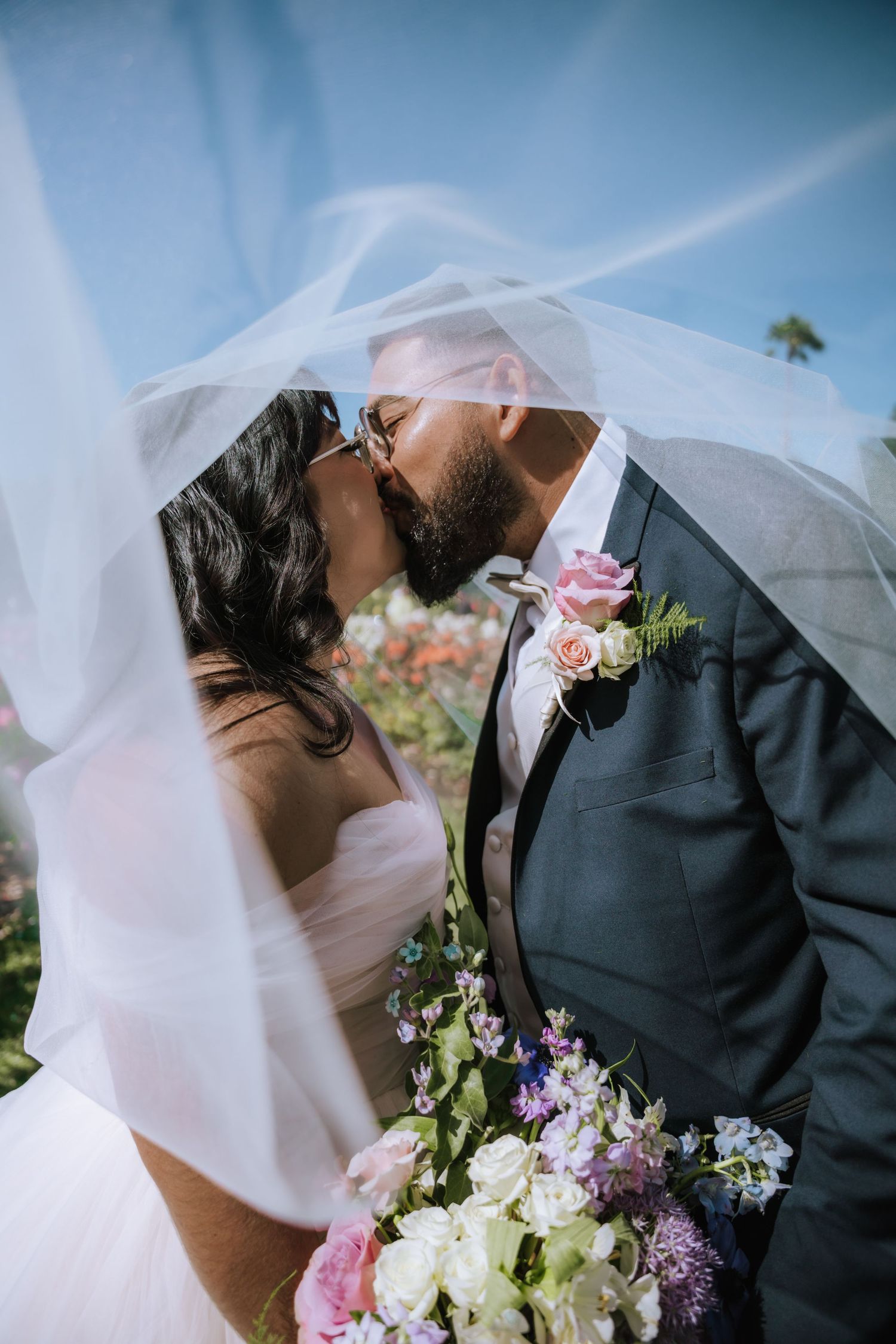 A couple shares a romantic kiss under a flowing white veil with colorful flowers and palm trees in the background.