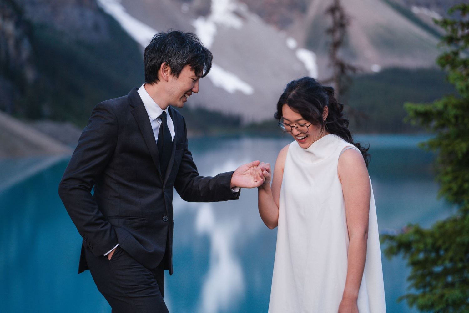 A couple shares a candid moment by a turquoise mountain lake, with snow-capped peaks in the background.